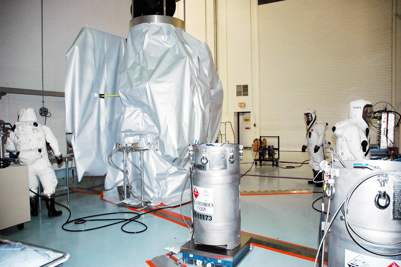 KENNEDY SPACE CENTER, FLA. -  Workers at Astrotech Space Operations in Titusville, Fla., begin fueling operations of the Deep Impact spacecraft, seen wrapped in a protective cover in the background. Scheduled for liftoff Jan. 12,  Deep Impact will probe beneath the surface of Comet Tempel 1 on July 4, 2005, when the comet is 83 million miles from Earth, and reveal the secrets of its interior.   After releasing a 3- by 3-foot projectile to crash onto the surface, Deep Impact’s flyby spacecraft will collect pictures and data of how the  crater forms, measuring the crater’s depth and diameter, as well as the composition of the interior of the crater and any material thrown out, and determining the changes in natural outgassing produced by the impact.  It will send the data back to Earth through the antennas of the Deep Space Network.  Deep Impact is a NASA Discovery mission.