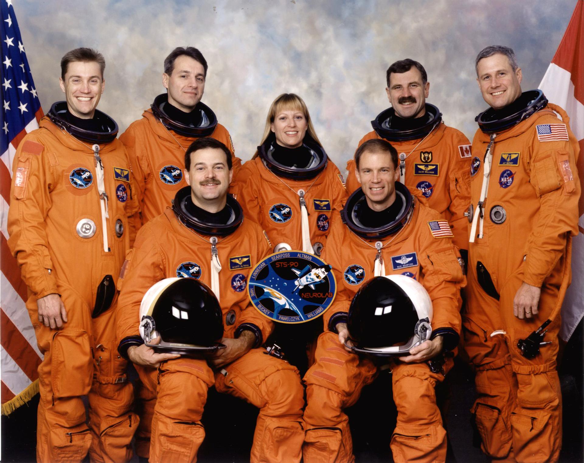 Seven astronauts pose for crew photo in orange spacesuits in front of flags.