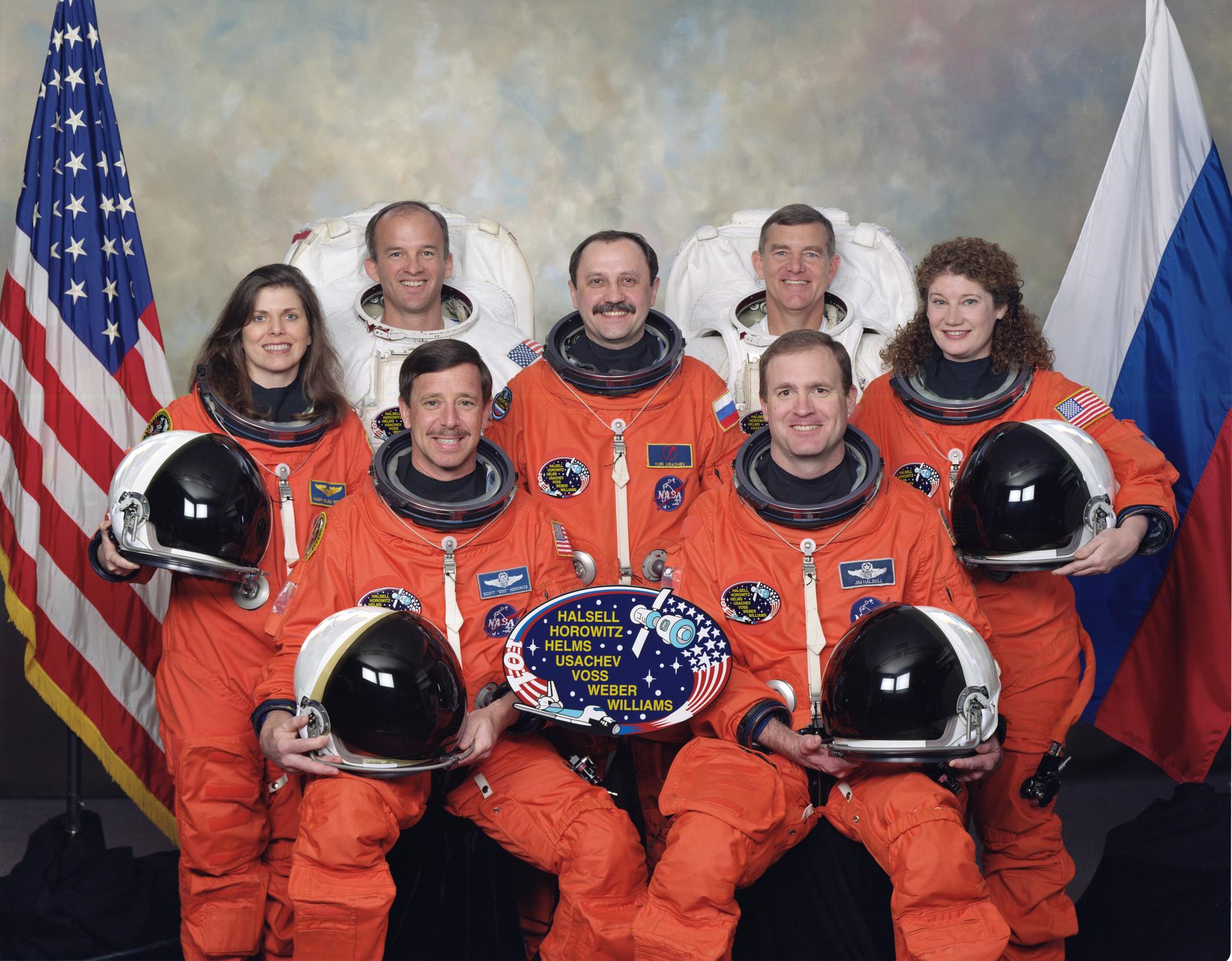 Seven astronauts in orange and white spacesuits pose for portrait in front of flags