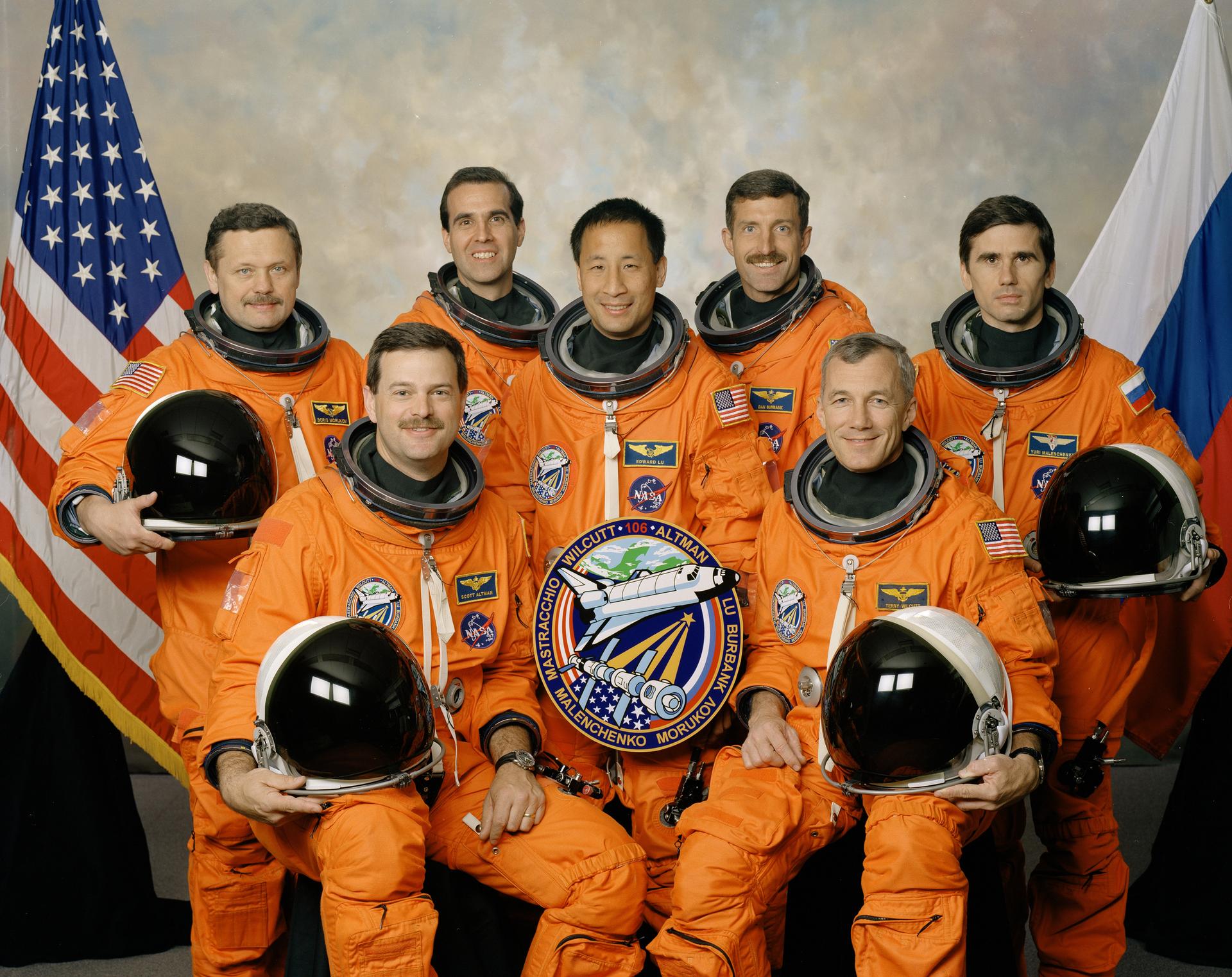 Portrait of STS-106 crew in orange space suites holding helmets in front of US and Russian flags.