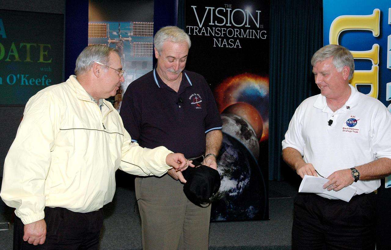 KENNEDY SPACE CENTER, FLA.  -  NASA Administrator Sean O’Keefe (center) is presented with a Deep Impact hat in the Press Site Auditorium following his report to employees on the state of the Agency.  He is accompanied on stage by Center Director Jim Kennedy (right). The update was broadcast live via NASA Television. O'Keefe focused on the achievements of 2004 and the goals set for 2005.  His remarks emphasized the milestones met in NASA's Vision for Space Exploration, including the launch of the comet-chasing Deep Impact mission and the landing of the Huygens probe on Jupiter’s moon Titan, both occurring in the past two days, and the progress made in meeting the requirements to return the Space Shuttle to flight.  O’Keefe’s briefing included a dialogue with Associate Administrator of NASA’s Office of Exploration Systems Craig Steidle and Center Director Jim Kennedy, live; and Manager of the Space Station Office Bill Gerstenmaier and Director of Advanced Planning and Jet Propulsion Laboratory Charles Elachi, via satellite.