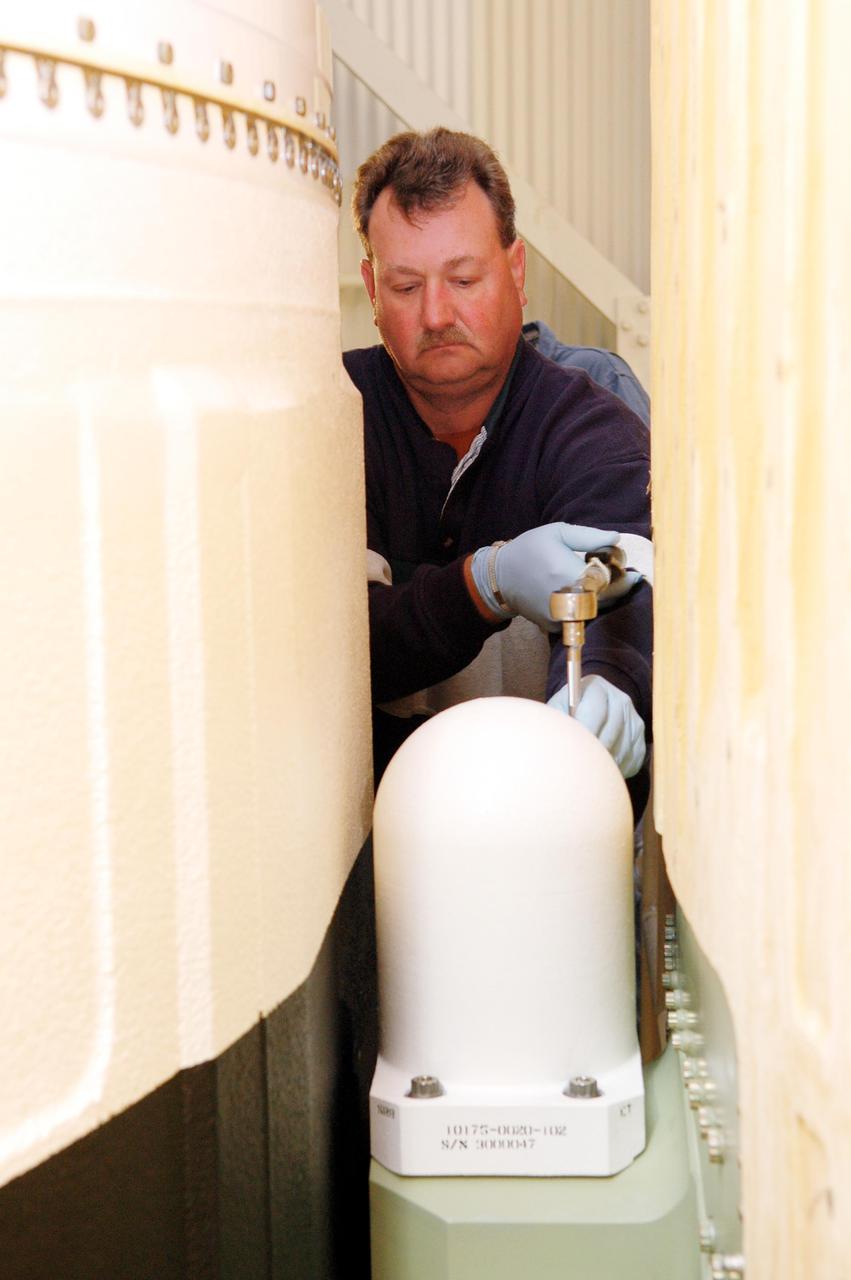 KENNEDY SPACE CENTER, FLA. - In the Vehicle Assembly Building, Lead Technician Todd Reeves, with United Space Alliance, attaches one of two bolt catchers on orbiter Discovery’s External Tank. A bolt catcher is a vertical bolt mechanism at the forward end of the External Tank that attaches each booster to the tank. At approximately two minutes into launch, SRB separation begins when pyrotechnic devices fire to break the 25-inch, 62-pound steel bolts. One half of the bolt is caught in canister-like 'bolt catchers' located on the tank; the other half remains with the boosters. Discovery is flying with a modified bolt catcher, which was upgraded from a two-piece welded design to a one-piece, machine-made design as part of NASA's effort to return to safe, reliable spaceflight. Eliminating the weld makes a structurally stronger bolt catcher design. Though the bolt catcher is mounted on the External Tank, it is considered part of the Solid Rocket Booster element design. It is built by Summa Technologies, Inc. in Huntsville, Ala., insulated at Lockheed Martin’s Michoud Assembly Facility in New Orleans, and installed on the External Tank at KSC.