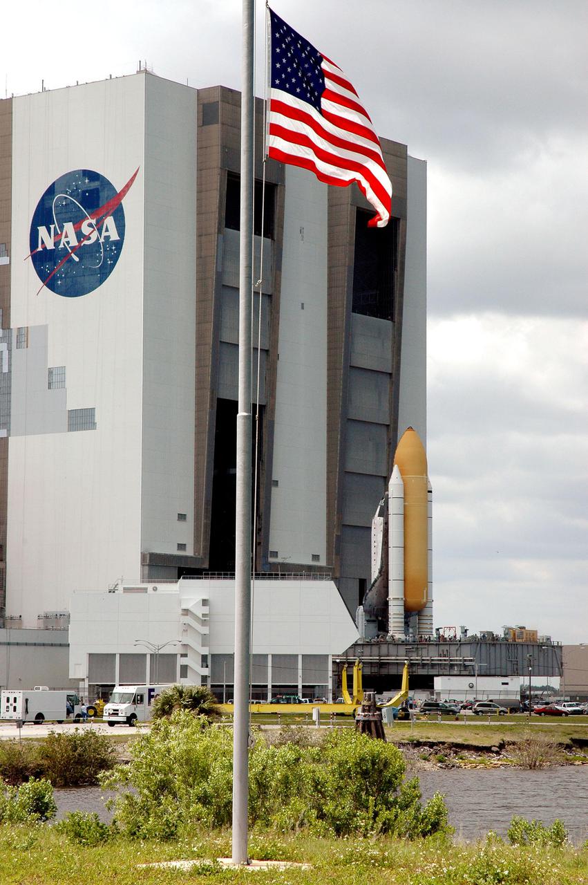 KENNEDY SPACE CENTER, FLA. -  —  Looking from the Press Site across the Turn Basin at NASA’s Kennedy Space Center, the American flag appears to salute Space Shuttle Discovery as she marks a major milestone for Return to Flight, rolling out to Launch Pad 39B.  First motion out of the Vehicle Assembly Building, behind the flag, was at 2:04 p.m. EDT. The Shuttle sits atop the Mobile Launcher Platform, which is moved by the Crawler-Transporter underneath.  The Crawler is 20 feet high, 131 feet long and 114 feet wide.  It moves on eight tracks, each containing 57 shoes, or cleats, weighing one ton each.  Loaded with the Space Shuttle, the Crawler can move at a maximum speed of approximately 1 mile an hour. A leveling system in the Crawler keeps the Shuttle vertical while negotiating the 5 percent grade leading to the top of the launch pad.  Launch of Discovery on its Return to Flight mission, STS-114, is targeted for May 15 with a launch window that extends to June 3. During its 12-day mission, Discovery’s seven-person crew will test new hardware and techniques to improve Shuttle safety, as well as deliver supplies to the International Space Station.  Discovery was moved on March 29 from the Orbiter Processing Facility to the VAB and attached to its propulsion elements, a redesigned ET and twin SRBs.