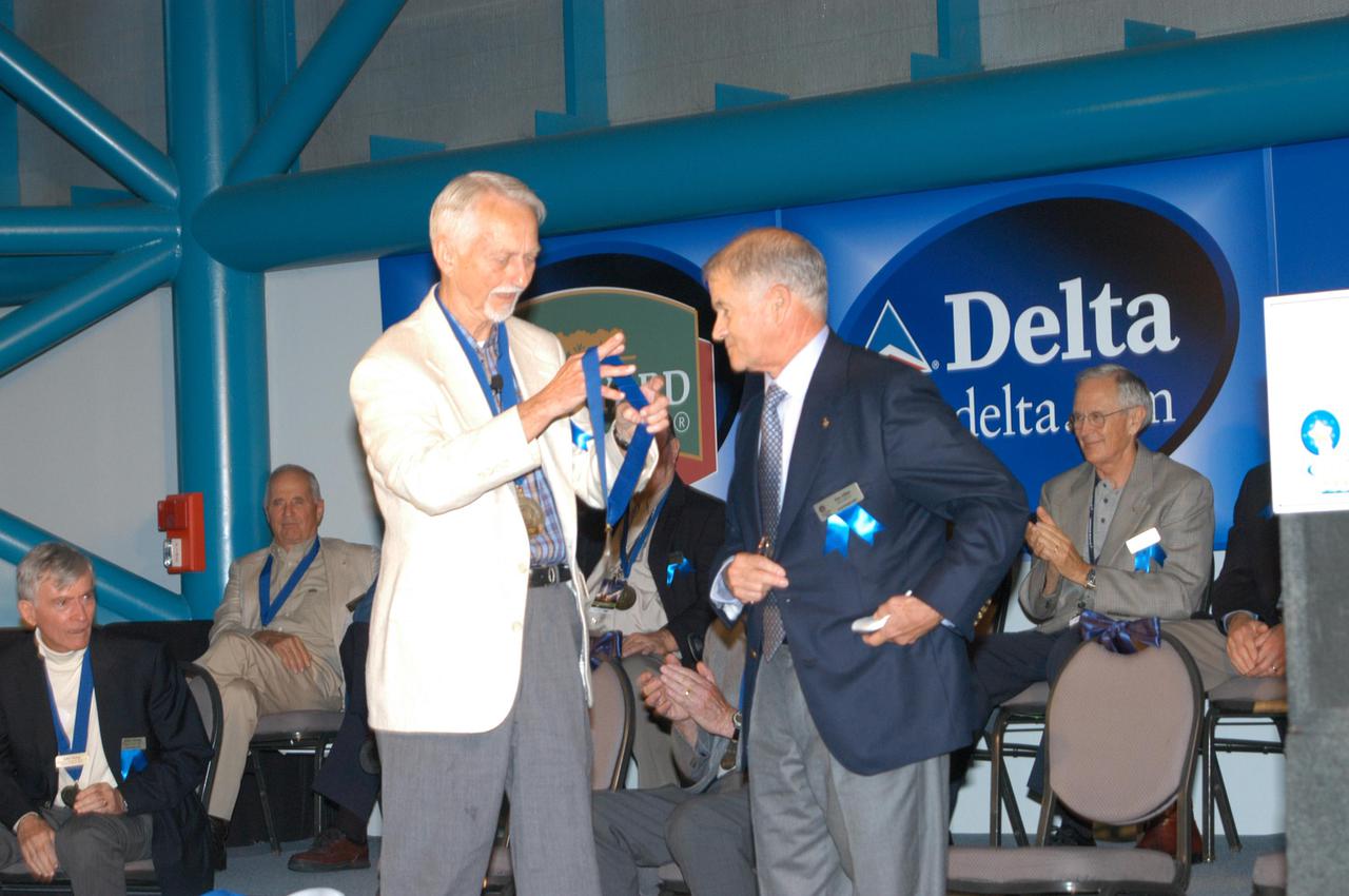 KENNEDY SPACE CENTER, FLA. -  At the Astronaut Hall of Fame Induction Ceremony, Chairman Owen Garriott prepares to place a medal on the neck of new inductee Joe Allen.  Current Hall of Famers in the background are John Young and Dick Gordon (at left) and Al Worden (at right).  The other new inductees are Bruce McCandless and Gordon Fullerton.  The event is being held in the Kennedy Space Center Visitor Complex’s Apollo_Saturn V Center. Recognized for their individual flight accomplishments and contributions to the success and future success of the U.S. space program, this elite group of inductees is among only 60 astronauts to be honored in the Hall of Fame and the fourth group of Space Shuttle astronauts named.