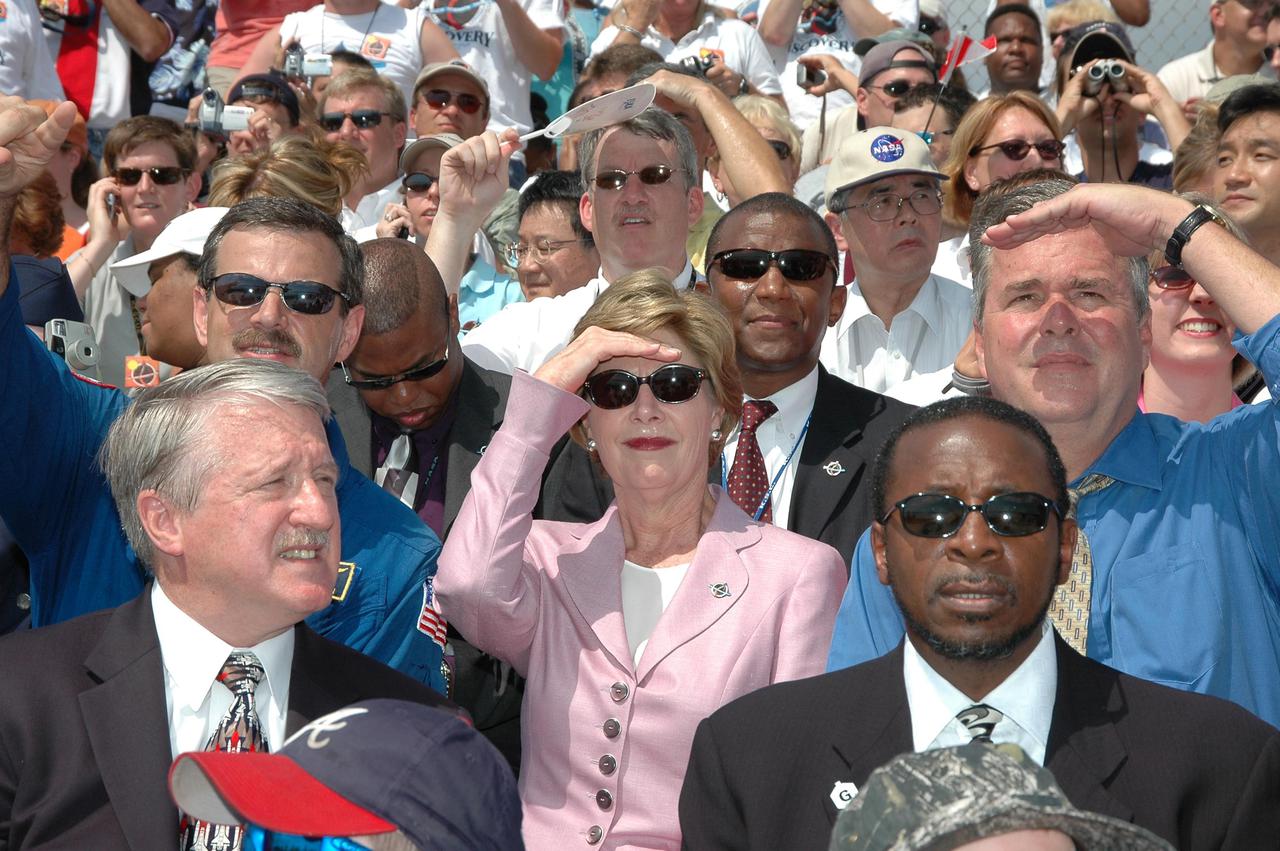 KENNEDY SPACE CENTER, FLA. - In the stands at NASA Kennedy Space Center’s Banana Creek viewing site, First Lady Laura Bush (center) watches Launch Pad 39B for the liftoff of Space Shuttle Discovery on Return to Flight mission STS-114, scheduled to lift off at 10:39 a.m. EDT. She is flanked by astronaut Scott Altmann at left and Florida Gov. Jeb Bush at right. In front of her are Michael O’Brien (left), assistant administrator for External Relations, and Woodrow Whitlow Jr. (right), KSC deputy director. Mrs. Bush is only the third First Lady to witness a Space Shuttle launch at KSC. On this mission to the International Space Station the crew will perform inspections on-orbit for the first time of all of the Reinforced Carbon-Carbon (RCC) panels on the leading edge of the wings and the Thermal Protection System tiles using the new Canadian-built Orbiter Boom Sensor System and the data from 176 impact and temperature sensors. Mission Specialists will also practice repair techniques on RCC and tile samples during a spacewalk in the payload bay. During two additional spacewalks, the crew will install the External Stowage Platform-2, equipped with spare part assemblies, and a replacement Control Moment Gyroscope contained in the Lightweight Multi-Purpose Experiment Support Structure. The 12-day mission is expected to end with touchdown at the Shuttle Landing Facility on Aug. 7.