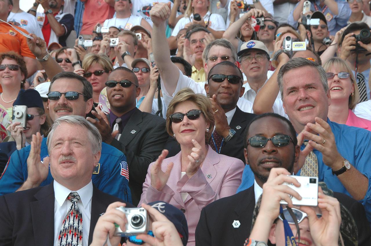 KENNEDY SPACE CENTER, FLA. -   In the stands at NASA Kennedy Space Center’s  Banana Creek viewing site, First Lady Laura Bush (center) applauds the successful liftoff of Space Shuttle Discovery on Return to Flight mission STS-114 at 10:39 a.m. EDT from Launch Pad 39B.  She is flanked by astronaut Scott Altmann at left and Florida Gov. Jeb Bush at right.  KSC Deputy Director Woodrow Whitlow Jr. is in front of the governor.  On this mission to the International Space Station the crew will perform inspections on-orbit for the first time of all of the Reinforced Carbon-Carbon (RCC) panels on the leading edge of the wings and the Thermal Protection System tiles using the new Canadian-built Orbiter Boom Sensor System and the data from 176 impact and temperature sensors. Mission Specialists will also practice repair techniques on RCC and tile samples during a spacewalk in the payload bay.  During two additional spacewalks, the crew will install the External Stowage Platform-2, equipped with spare part assemblies, and a replacement Control Moment Gyroscope contained in the Lightweight Multi-Purpose Experiment Support Structure. The 12-day mission is expected to end with touchdown at the Shuttle Landing Facility on Aug. 7.
