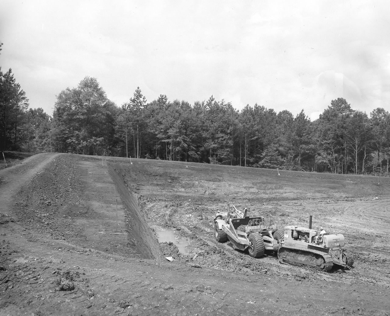 At its founding, the Marshall Space Flight Center (MSFC) inherited the Army’s Jupiter and Redstone test stands, but much larger facilities were needed for the giant stages of the Saturn V. From 1960 to 1964, the existing stands were remodeled and a sizable new test area was developed. The new comprehensive test complex for propulsion and structural dynamics was unique within the nation and the free world, and they remain so today because they were constructed with foresight to meet the future as well as on going needs. Construction of the S-IC Static test stand complex began in 1961 in the west test area of MSFC, and was completed in 1964. The S-IC static test stand was designed to develop and test the 138-ft long and 33-ft diameter Saturn V S-IC first stage, or booster stage, weighing in at 280,000 pounds. Required to hold down the brute force of a 7,500,000-pound thrust produced by 5 F-1 engines, the S-IC static test stand was designed and constructed with the strength of hundreds of tons of steel and 12,000,000 pounds of cement, planted down to bedrock 40 feet below ground level. The foundation walls, constructed with concrete and steel, are 4 feet thick. The base structure consists of four towers with 40-foot-thick walls extending upward 144 feet above ground level. The structure was topped by a crane with a 135-foot boom. With the boom in the upright position, the stand was given an overall height of 405 feet, placing it among the highest structures in Alabama at the time.  In this photo, taken July 13, 1961, progress is made with the excavation and preparation of the S-IC test stand site.  