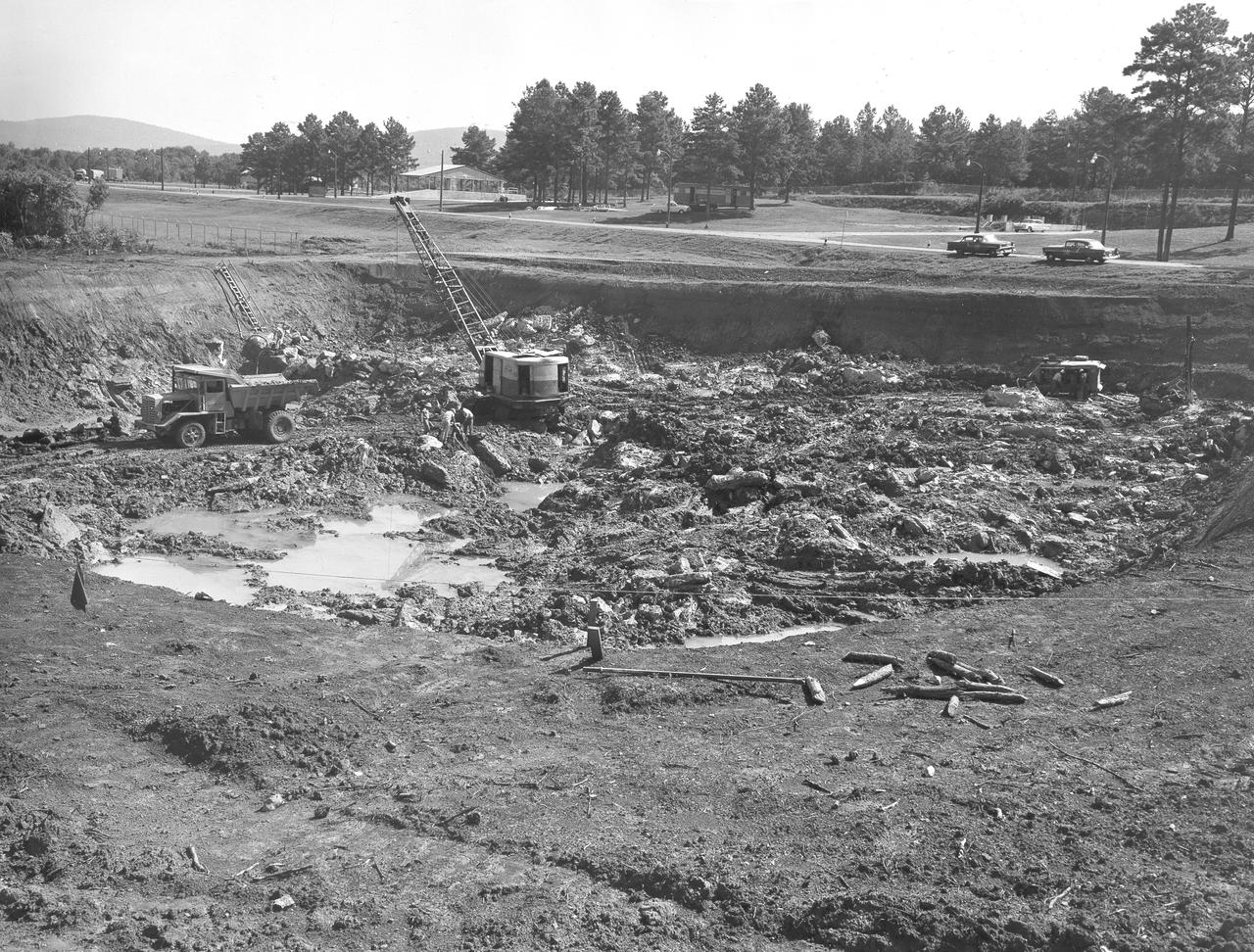 At its founding, the Marshall Space Flight Center (MSFC) inherited the Army’s Jupiter and Redstone test stands, but much larger facilities were needed for the giant stages of the Saturn V. From 1960 to 1964, the existing stands were remodeled and a sizable new test area was developed. The new comprehensive test complex for propulsion and structural dynamics was unique within the nation and the free world, and they remain so today because they were constructed with foresight to meet the future as well as on going needs. Construction of the S-IC Static test stand complex began in 1961 in the west test area of MSFC, and was completed in 1964. The S-IC static test stand was designed to develop and test the 138-ft long and 33-ft diameter Saturn V S-IC first stage, or booster stage, weighing in at 280,000 pounds. Required to hold down the brute force of a 7,500,000-pound thrust produced by 5 F-1 engines, the S-IC static test stand was designed and constructed with the strength of hundreds of tons of steel and 12,000,000 pounds of cement, planted down to bedrock 40 feet below ground level. The foundation walls, constructed with concrete and steel, are 4 feet thick. The base structure consists of four towers with 40-foot-thick walls extending upward 144 feet above ground level. The structure was topped by a crane with a 135-foot boom. With the boom in the upright position, the stand was given an overall height of 405 feet, placing it among the highest structures in Alabama at the time. In this photo, taken July 31, 1961, work is continued in the clearing of the test stand site.