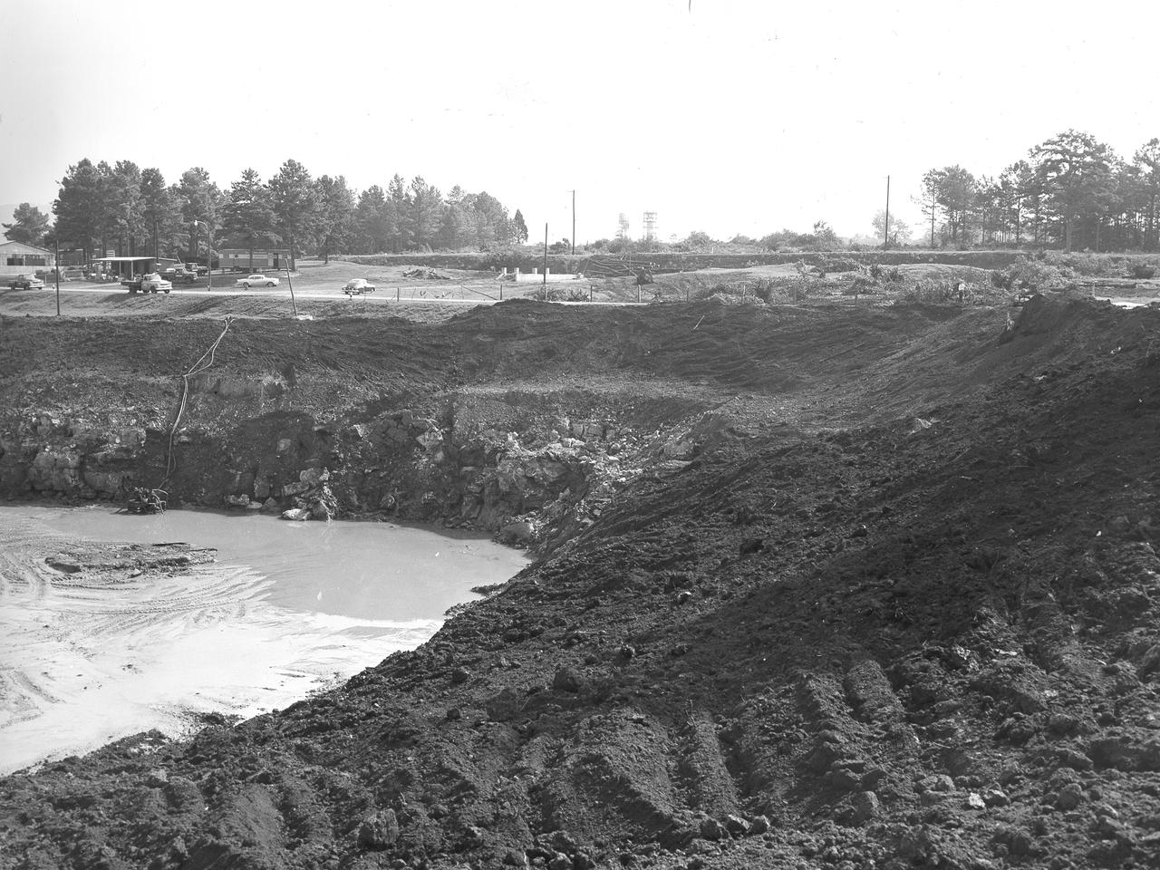 At its founding, the Marshall Space Flight Center (MSFC) inherited the Army’s Jupiter and Redstone test stands, but much larger facilities were needed for the giant stages of the Saturn V. From 1960 to 1964, the existing stands were remodeled and a sizable new test area was developed. The new comprehensive test complex for propulsion and structural dynamics was unique within the nation and the free world, and they remain so today because they were constructed with foresight to meet the future as well as on going needs. Construction of the S-IC Static test stand complex began in 1961 in the west test area of MSFC, and was completed in 1964. The S-IC static test stand was designed to develop and test the 138-ft long and 33-ft diameter Saturn V S-IC first stage, or booster stage, weighing in at 280,000 pounds. Required to hold down the brute force of a 7,500,000-pound thrust produced by 5 F-1 engines, the S-IC static test stand was designed and constructed with the strength of hundreds of tons of steel and 12,000,000 pounds of cement, planted down to bedrock 40 feet below ground level. The foundation walls, constructed with concrete and steel, are 4 feet thick. The base structure consists of four towers with 40-foot-thick walls extending upward 144 feet above ground level. The structure was topped by a crane with a 135-foot boom. With the boom in the upright position, the stand was given an overall height of 405 feet, placing it among the highest structures in Alabama at the time. This photo shows the construction progress of the test stand as of August 18, 1961.