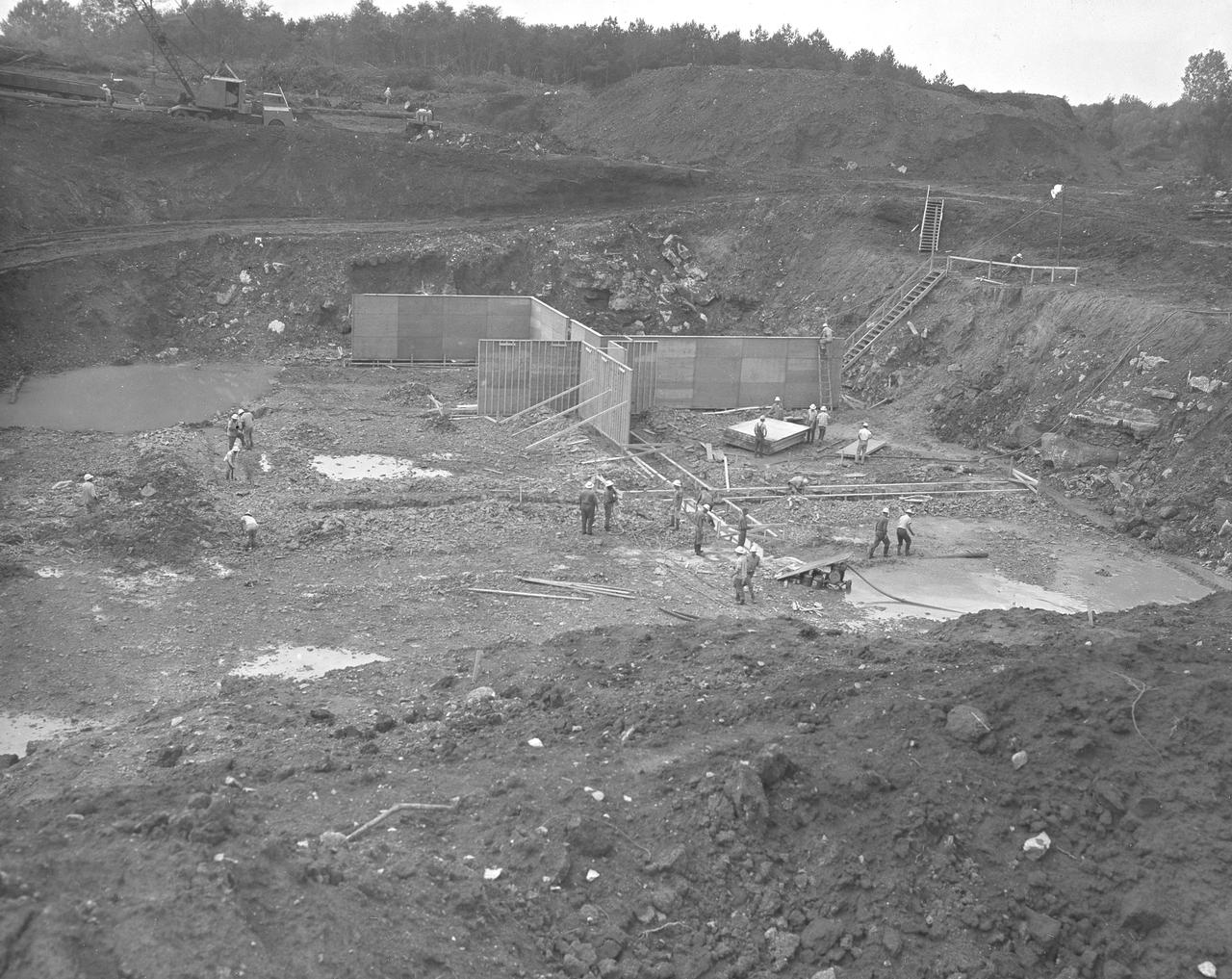 At its founding, the Marshall Space Flight Center (MSFC) inherited the Army’s Jupiter and Redstone test stands, but much larger facilities were needed for the giant stages of the Saturn V. From 1960 to 1964, the existing stands were remodeled and a sizable new test area was developed. The new comprehensive test complex for propulsion and structural dynamics was unique within the nation and the free world, and they remain so today because they were constructed with foresight to meet the future as well as on going needs. Construction of the S-IC Static test stand complex began in 1961 in the west test area of MSFC, and was completed in 1964. The S-IC static test stand was designed to develop and test the 138-ft long and 33-ft diameter Saturn V S-IC first stage, or booster stage, weighing in at 280,000 pounds. Required to hold down the brute force of a 7,500,000-pound thrust produced by 5 F-1 engines, the S-IC static test stand was designed and constructed with the strength of hundreds of tons of steel and 12,000,000 pounds of cement, planted down to bedrock 40 feet below ground level. The foundation walls, constructed with concrete and steel, are 4 feet thick. The base structure consists of four towers with 40-foot-thick walls extending upward 144 feet above ground level. The structure was topped by a crane with a 135-foot boom. With the boom in the upright position, the stand was given an overall height of 405 feet, placing it among the highest structures in Alabama at the time. This photo, taken September 1, 1961, shows the construction of forms which became the concrete foundation for the massive stand.