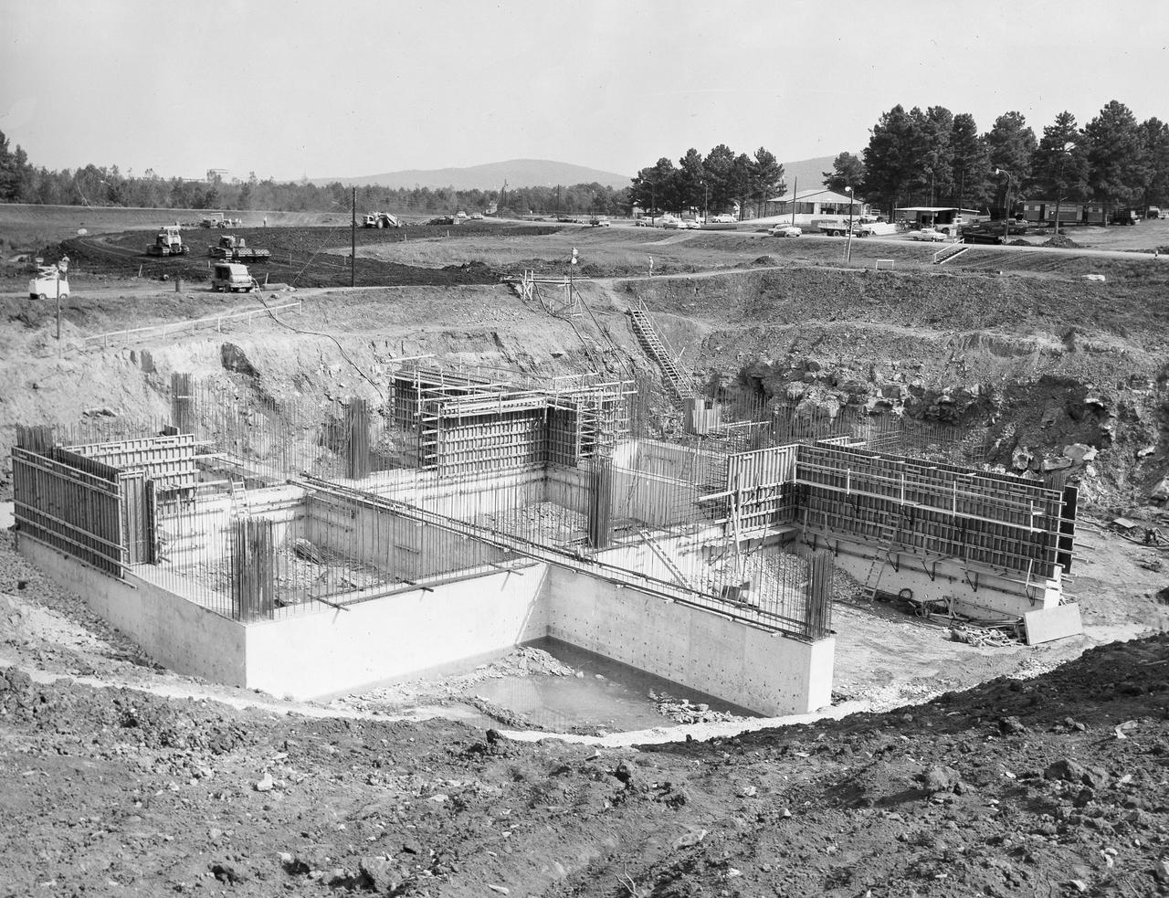 At its founding, the Marshall Space Flight Center (MSFC) inherited the Army’s Jupiter and Redstone test stands, but much larger facilities were needed for the giant stages of the Saturn V. From 1960 to 1964, the existing stands were remodeled and a sizable new test area was developed. The new comprehensive test complex for propulsion and structural dynamics was unique within the nation and the free world, and they remain so today because they were constructed with foresight to meet the future as well as on going needs. Construction of the S-IC Static test stand complex began in 1961 in the west test area of MSFC, and was completed in 1964. The S-IC static test stand was designed to develop and test the 138-ft long and 33-ft diameter Saturn V S-IC first stage, or booster stage, weighing in at 280,000 pounds. Required to hold down the brute force of a 7,500,000-pound thrust produced by 5 F-1 engines, the S-IC static test stand was designed and constructed with the strength of hundreds of tons of steel and 12,000,000 pounds of cement, planted down to bedrock 40 feet below ground level. The foundation walls, constructed with concrete and steel, are 4 feet thick. The base structure consists of four towers with 40-foot-thick walls extending upward 144 feet above ground level. The structure was topped by a crane with a 135-foot boom. With the boom in the upright position, the stand was given an overall height of 405 feet, placing it among the highest structures in Alabama at the time. This photo, taken September 29, 1961, shows the progress of the concrete walls for the stand’s foundation. Some of the walls have been poured and some of the concrete forms have been removed.