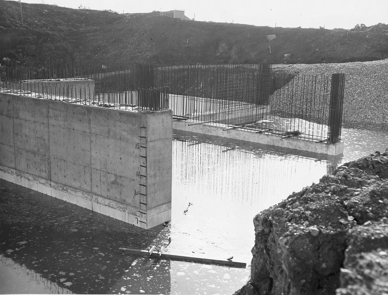 At its founding, the Marshall Space Flight Center (MSFC) inherited the Army’s Jupiter and Redstone test stands, but much larger facilities were needed for the giant stages of the Saturn V. From 1960 to 1964, the existing stands were remodeled and a sizable new test area was developed. The new comprehensive test complex for propulsion and structural dynamics was unique within the nation and the free world, and they remain so today because they were constructed with foresight to meet the future as well as on going needs. Construction of the S-IC Static test stand complex began in 1961 in the west test area of MSFC, and was completed in 1964. The S-IC static test stand was designed to develop and test the 138-ft long and 33-ft diameter Saturn V S-IC first stage, or booster stage, weighing in at 280,000 pounds. Required to hold down the brute force of a 7,500,000-pound thrust produced by 5 F-1 engines, the S-IC static test stand was designed and constructed with the strength of hundreds of tons of steel and 12,000,000 pounds of cement, planted down to bedrock 40 feet below ground level. The foundation walls, constructed with concrete and steel, are 4 feet thick. The base structure consists of four towers with 40-foot-thick walls extending upward 144 feet above ground level. The structure was topped by a crane with a 135-foot boom. With the boom in the upright position, the stand was given an overall height of 405 feet, placing it among the highest structures in Alabama at the time. Construction of the S-IC test stand came to a halt at the end of September as the determination was made that the Saturn booster size had to be increased. As a result, the stand had to be modified. With construction delayed, and pumps turned off, this photo, taken December 1, 1961, shows the abandoned site with floods at the 6 ft mark. The flooding was caused by the disturbance of a natural spring months prior during the excavation of the site. 