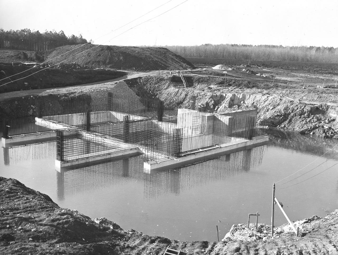 At its founding, the Marshall Space Flight Center (MSFC) inherited the Army’s Jupiter and Redstone test stands, but much larger facilities were needed for the giant stages of the Saturn V. From 1960 to 1964, the existing stands were remodeled and a sizable new test area was developed. The new comprehensive test complex for propulsion and structural dynamics was unique within the nation and the free world, and they remain so today because they were constructed with foresight to meet the future as well as on going needs. Construction of the S-IC Static test stand complex began in 1961 in the west test area of MSFC, and was completed in 1964. The S-IC static test stand was designed to develop and test the 138-ft long and 33-ft diameter Saturn V S-IC first stage, or booster stage, weighing in at 280,000 pounds. Required to hold down the brute force of a 7,500,000-pound thrust produced by 5 F-1 engines, the S-IC static test stand was designed and constructed with the strength of hundreds of tons of steel and 12,000,000 pounds of cement, planted down to bedrock 40 feet below ground level. The foundation walls, constructed with concrete and steel, are 4 feet thick. The base structure consists of four towers with 40-foot-thick walls extending upward 144 feet above ground level. The structure was topped by a crane with a 135-foot boom. With the boom in the upright position, the stand was given an overall height of 405 feet, placing it among the highest structures in Alabama at the time. Construction of the S-IC test stand came to a halt at the end of September as the determination was made that the Saturn booster size had to be increased. As a result, the stand had to be modified. With construction delayed, and pumps turned off, this photo, taken December 1, 1961, shows the abandoned site with floods at the 6 ft mark. The flooding was caused by the disturbance of a natural spring months prior during the excavation of the site. 