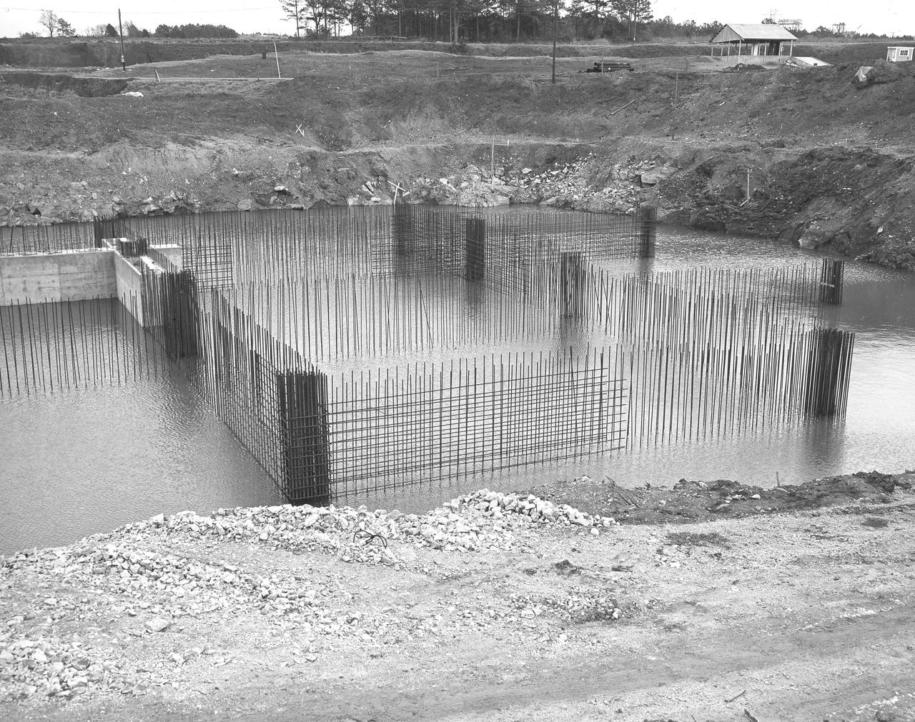 At its founding, the Marshall Space Flight Center (MSFC) inherited the Army’s Jupiter and Redstone test stands, but much larger facilities were needed for the giant stages of the Saturn V. From 1960 to 1964, the existing stands were remodeled and a sizable new test area was developed. The new comprehensive test complex for propulsion and structural dynamics was unique within the nation and the free world, and they remain so today because they were constructed with foresight to meet the future as well as on going needs. Construction of the S-IC Static test stand complex began in 1961 in the west test area of MSFC, and was completed in 1964. The S-IC static test stand was designed to develop and test the 138-ft long and 33-ft diameter Saturn V S-IC first stage, or booster stage, weighing in at 280,000 pounds. Required to hold down the brute force of a 7,500,000-pound thrust produced by 5 F-1 engines, the S-IC static test stand was designed and constructed with the strength of hundreds of tons of steel and 12,000,000 pounds of cement, planted down to bedrock 40 feet below ground level. The foundation walls, constructed with concrete and steel, are 4 feet thick. The base structure consists of four towers with 40-foot-thick walls extending upward 144 feet above ground level. The structure was topped by a crane with a 135-foot boom. With the boom in the upright position, the stand was given an overall height of 405 feet, placing it among the highest structures in Alabama at the time. Construction of the S-IC test stand came to a halt at the end of September as the determination was made that the Saturn booster size had to be increased. As a result, the stand had to be modified. With construction delayed, and pumps turned off, this photo, taken December 4, 1961, shows the abandoned site with floods at the 11 ft mark. The flooding was caused by the disturbance of a natural spring months prior during the excavation of the site. 