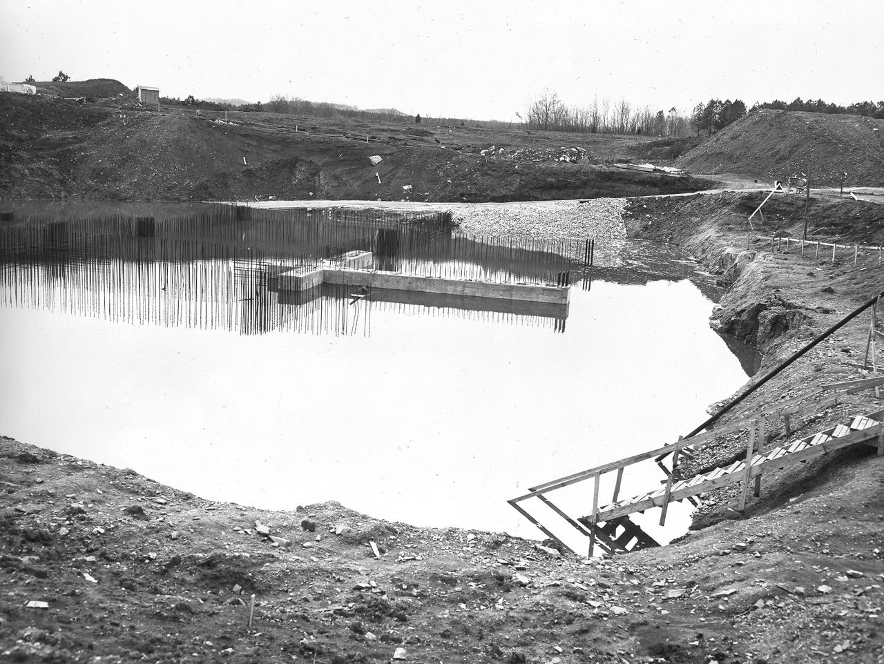 At its founding, the Marshall Space Flight Center (MSFC) inherited the Army’s Jupiter and Redstone test stands, but much larger facilities were needed for the giant stages of the Saturn V. From 1960 to 1964, the existing stands were remodeled and a sizable new test area was developed. The new comprehensive test complex for propulsion and structural dynamics was unique within the nation and the free world, and they remain so today because they were constructed with foresight to meet the future as well as on going needs. Construction of the S-IC Static test stand complex began in 1961 in the west test area of MSFC, and was completed in 1964. The S-IC static test stand was designed to develop and test the 138-ft long and 33-ft diameter Saturn V S-IC first stage, or booster stage, weighing in at 280,000 pounds. Required to hold down the brute force of a 7,500,000-pound thrust produced by 5 F-1 engines, the S-IC static test stand was designed and constructed with the strength of hundreds of tons of steel and 12,000,000 pounds of cement, planted down to bedrock 40 feet below ground level. The foundation walls, constructed with concrete and steel, are 4 feet thick. The base structure consists of four towers with 40-foot-thick walls extending upward 144 feet above ground level. The structure was topped by a crane with a 135-foot boom. With the boom in the upright position, the stand was given an overall height of 405 feet, placing it among the highest structures in Alabama at the time. Construction of the S-IC test stand came to a halt at the end of September as the determination was made that the Saturn booster size had to be increased. As a result, the stand had to be modified. With construction delayed, and pumps turned off, this photo, taken December 8, 1961, shows the abandoned site with floods at the 16 ft mark. The flooding was caused by the disturbance of a natural spring months prior during the excavation of the site.     