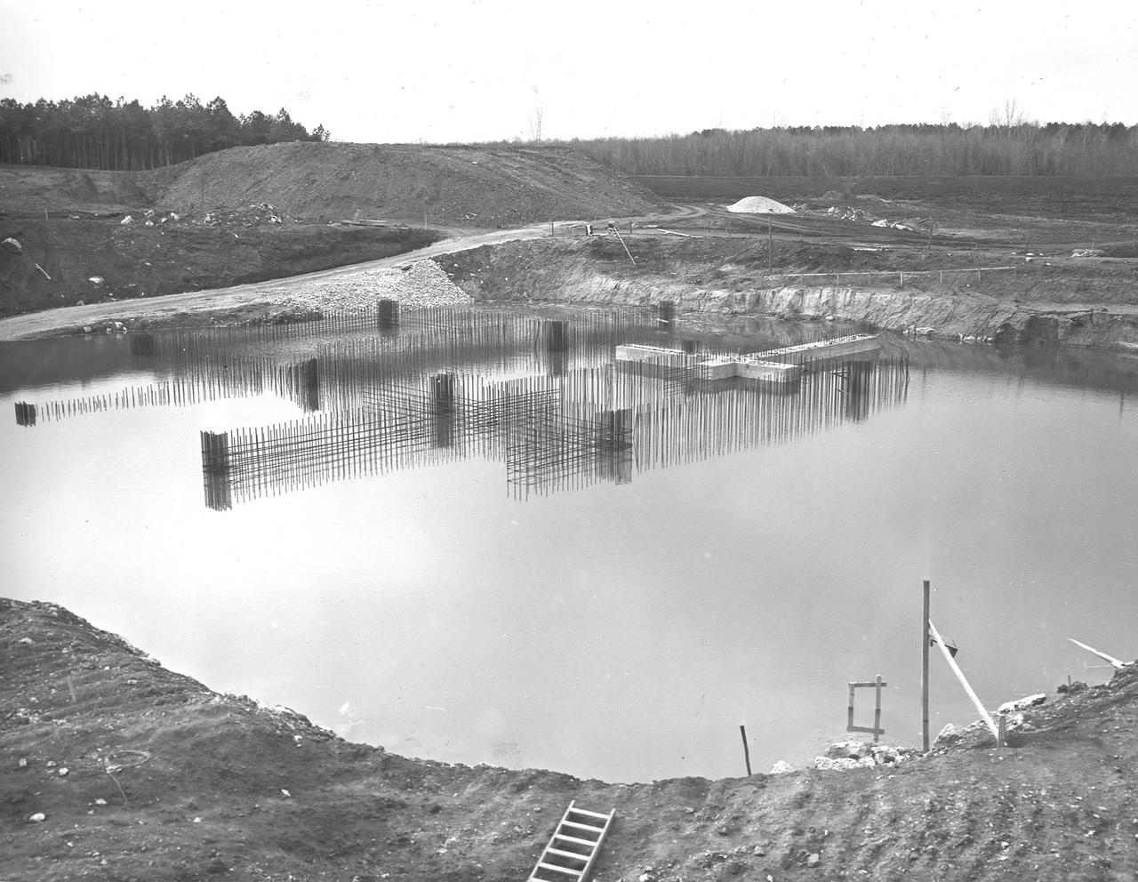 At its founding, the Marshall Space Flight Center (MSFC) inherited the Army’s Jupiter and Redstone test stands, but much larger facilities were needed for the giant stages of the Saturn V. From 1960 to 1964, the existing stands were remodeled and a sizable new test area was developed. The new comprehensive test complex for propulsion and structural dynamics was unique within the nation and the free world, and they remain so today because they were constructed with foresight to meet the future as well as on going needs. Construction of the S-IC Static test stand complex began in 1961 in the west test area of MSFC, and was completed in 1964. The S-IC static test stand was designed to develop and test the 138-ft long and 33-ft diameter Saturn V S-IC first stage, or booster stage, weighing in at 280,000 pounds. Required to hold down the brute force of a 7,500,000-pound thrust produced by 5 F-1 engines, the S-IC static test stand was designed and constructed with the strength of hundreds of tons of steel and 12,000,000 pounds of cement, planted down to bedrock 40 feet below ground level. The foundation walls, constructed with concrete and steel, are 4 feet thick. The base structure consists of four towers with 40-foot-thick walls extending upward 144 feet above ground level. The structure was topped by a crane with a 135-foot boom. With the boom in the upright position, the stand was given an overall height of 405 feet, placing it among the highest structures in Alabama at the time. Construction of the S-IC test stand came to a halt at the end of September as the determination was made that the Saturn booster size had to be increased. As a result, the stand had to be modified. With construction delayed, and pumps turned off, this photo, taken December 8, 1961, shows the abandoned site with floods at the 16 ft mark. The flooding was caused by the disturbance of a natural spring months prior during the excavation of the site. 