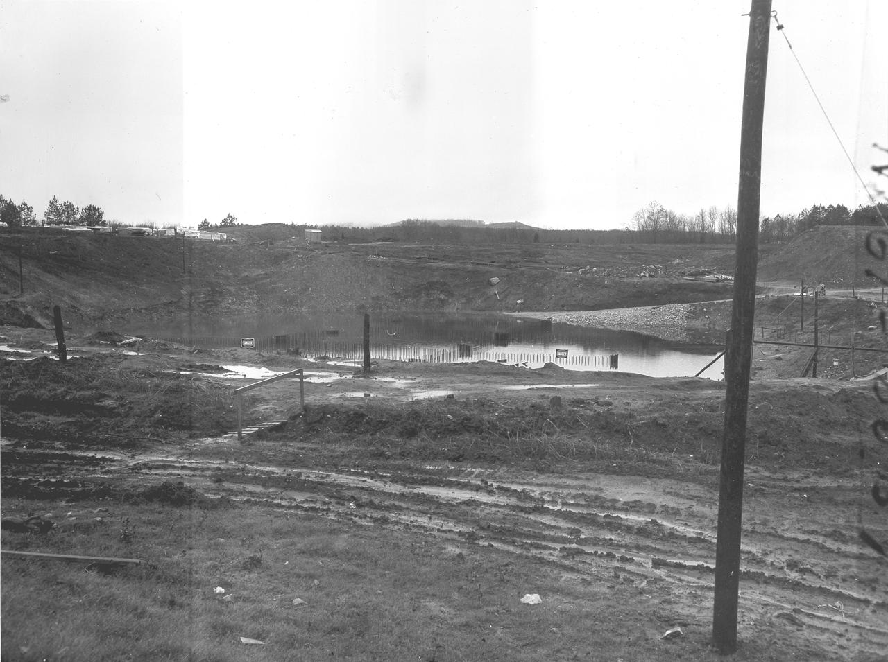 At its founding, the Marshall Space Flight Center (MSFC) inherited the Army’s Jupiter and Redstone test stands, but much larger facilities were needed for the giant stages of the Saturn V. From 1960 to 1964, the existing stands were remodeled and a sizable new test area was developed. The new comprehensive test complex for propulsion and structural dynamics was unique within the nation and the free world, and they remain so today because they were constructed with foresight to meet the future as well as on going needs. Construction of the S-IC Static test stand complex began in 1961 in the west test area of MSFC, and was completed in 1964. The S-IC static test stand was designed to develop and test the 138-ft long and 33-ft diameter Saturn V S-IC first stage, or booster stage, weighing in at 280,000 pounds. Required to hold down the brute force of a 7,500,000-pound thrust produced by 5 F-1 engines, the S-IC static test stand was designed and constructed with the strength of hundreds of tons of steel and 12,000,000 pounds of cement, planted down to bedrock 40 feet below ground level. The foundation walls, constructed with concrete and steel, are 4 feet thick. The base structure consists of four towers with 40-foot-thick walls extending upward 144 feet above ground level. The structure was topped by a crane with a 135-foot boom. With the boom in the upright position, the stand was given an overall height of 405 feet, placing it among the highest structures in Alabama at the time. Construction of the S-IC test stand came to a halt at the end of September as the determination was made that the Saturn booster size had to be increased. As a result, the stand had to be modified. With construction delayed, and pumps turned off, this photo, taken December 11, 1961, shows the abandoned site with floods above the 18 ft mark. The flooding was caused by the disturbance of a natural spring months prior during the excavation of the site. 