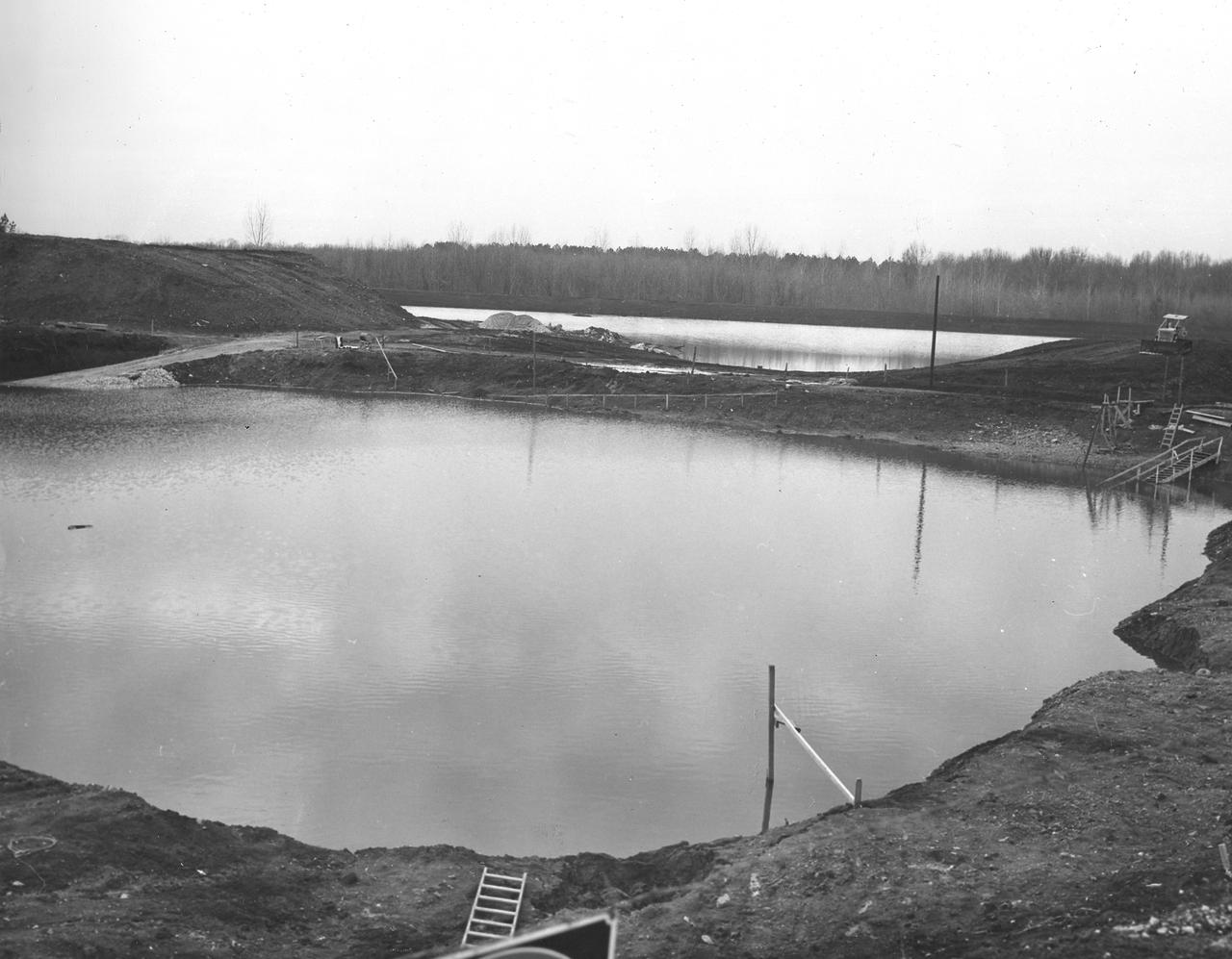 At its founding, the Marshall Space Flight Center (MSFC) inherited the Army’s Jupiter and Redstone test stands, but much larger facilities were needed for the giant stages of the Saturn V. From 1960 to 1964, the existing stands were remodeled and a sizable new test area was developed. The new comprehensive test complex for propulsion and structural dynamics was unique within the nation and the free world, and they remain so today because they were constructed with foresight to meet the future as well as on going needs. Construction of the S-IC Static test stand complex began in 1961 in the west test area of MSFC, and was completed in 1964. The S-IC static test stand was designed to develop and test the 138-ft long and 33-ft diameter Saturn V S-IC first stage, or booster stage, weighing in at 280,000 pounds. Required to hold down the brute force of a 7,500,000-pound thrust produced by 5 F-1 engines, the S-IC static test stand was designed and constructed with the strength of hundreds of tons of steel and 12,000,000 pounds of cement, planted down to bedrock 40 feet below ground level. The foundation walls, constructed with concrete and steel, are 4 feet thick. The base structure consists of four towers with 40-foot-thick walls extending upward 144 feet above ground level. The structure was topped by a crane with a 135-foot boom. With the boom in the upright position, the stand was given an overall height of 405 feet, placing it among the highest structures in Alabama at the time. Construction of the S-IC test stand came to a halt at the end of September as the determination was made that the Saturn booster size had to be increased. As a result, the stand had to be modified. With construction delayed, and pumps turned off, this photo, taken December 14, 1961, shows the abandoned site entirely flooded. The flooding was caused by the disturbance of a natural spring months prior during the excavation of the site. 