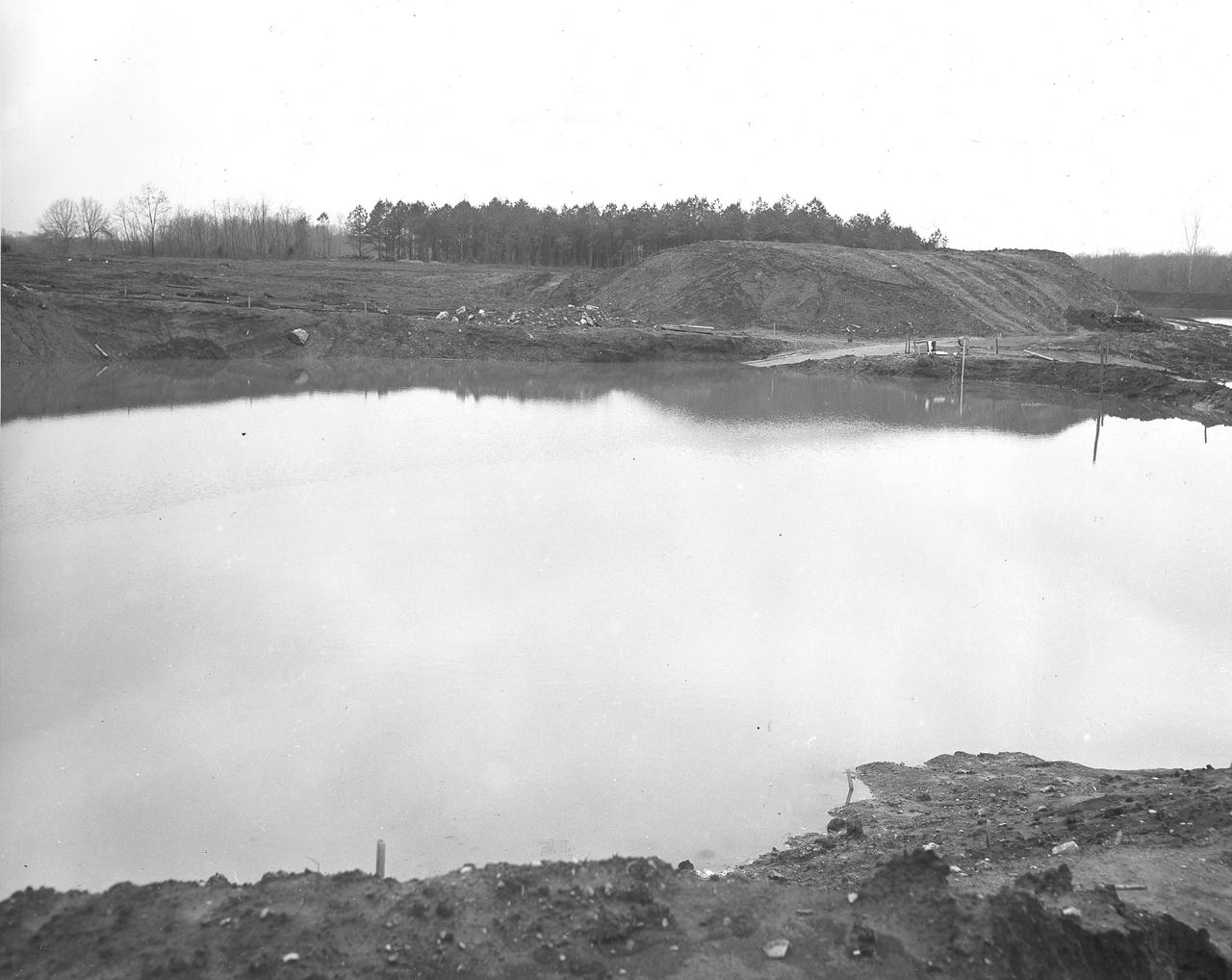 At its founding, the Marshall Space Flight Center (MSFC) inherited the Army’s Jupiter and Redstone test stands, but much larger facilities were needed for the giant stages of the Saturn V. From 1960 to 1964, the existing stands were remodeled and a sizable new test area was developed. The new comprehensive test complex for propulsion and structural dynamics was unique within the nation and the free world, and they remain so today because they were constructed with foresight to meet the future as well as on going needs. Construction of the S-IC Static test stand complex began in 1961 in the west test area of MSFC, and was completed in 1964. The S-IC static test stand was designed to develop and test the 138-ft long and 33-ft diameter Saturn V S-IC first stage, or booster stage, weighing in at 280,000 pounds. Required to hold down the brute force of a 7,500,000-pound thrust produced by 5 F-1 engines, the S-IC static test stand was designed and constructed with the strength of hundreds of tons of steel and 12,000,000 pounds of cement, planted down to bedrock 40 feet below ground level. The foundation walls, constructed with concrete and steel, are 4 feet thick. The base structure consists of four towers with 40-foot-thick walls extending upward 144 feet above ground level. The structure was topped by a crane with a 135-foot boom. With the boom in the upright position, the stand was given an overall height of 405 feet, placing it among the highest structures in Alabama at the time. Construction of the S-IC test stand came to a halt at the end of September as the determination was made that the Saturn booster size had to be increased. As a result, the stand had to be modified. With construction delayed, and pumps turned off, this photo, taken December 18, 1961, shows the abandoned site entirely flooded. The flooding was caused by the disturbance of a natural spring months prior during the excavation of the site. 