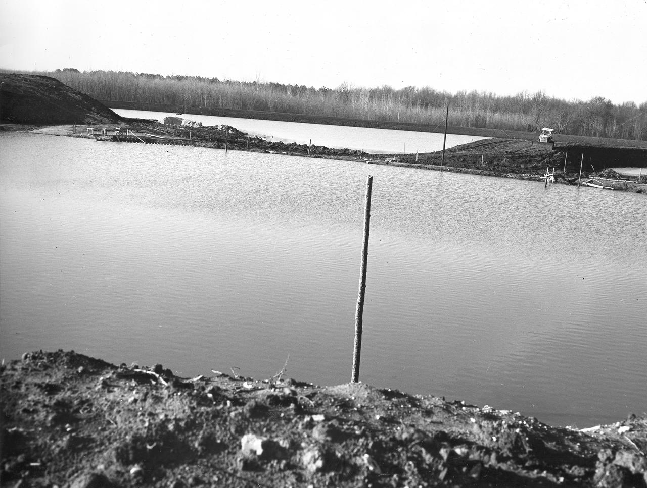 At its founding, the Marshall Space Flight Center (MSFC) inherited the Army’s Jupiter and Redstone test stands, but much larger facilities were needed for the giant stages of the Saturn V. From 1960 to 1964, the existing stands were remodeled and a sizable new test area was developed. The new comprehensive test complex for propulsion and structural dynamics was unique within the nation and the free world, and they remain so today because they were constructed with foresight to meet the future as well as on going needs. Construction of the S-IC Static test stand complex began in 1961 in the west test area of MSFC, and was completed in 1964. The S-IC static test stand was designed to develop and test the 138-ft long and 33-ft diameter Saturn V S-IC first stage, or booster stage, weighing in at 280,000 pounds. Required to hold down the brute force of a 7,500,000-pound thrust produced by 5 F-1 engines, the S-IC static test stand was designed and constructed with the strength of hundreds of tons of steel and 12,000,000 pounds of cement, planted down to bedrock 40 feet below ground level. The foundation walls, constructed with concrete and steel, are 4 feet thick. The base structure consists of four towers with 40-foot-thick walls extending upward 144 feet above ground level. The structure was topped by a crane with a 135-foot boom. With the boom in the upright position, the stand was given an overall height of 405 feet, placing it among the highest structures in Alabama at the time. Construction of the S-IC test stand came to a halt at the end of September as the determination was made that the Saturn booster size had to be increased. As a result, the stand had to be modified. With construction delayed, and pumps turned off, this photo, taken December 22, 1961, shows danger signs posted around the abandoned site with floods nearing the top. The flooding was caused by the disturbance of a natural spring months prior during the excavation of the site. 