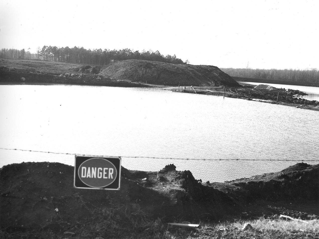 At its founding, the Marshall Space Flight Center (MSFC) inherited the Army’s Jupiter and Redstone test stands, but much larger facilities were needed for the giant stages of the Saturn V. From 1960 to 1964, the existing stands were remodeled and a sizable new test area was developed. The new comprehensive test complex for propulsion and structural dynamics was unique within the nation and the free world, and they remain so today because they were constructed with foresight to meet the future as well as on going needs. Construction of the S-IC Static test stand complex began in 1961 in the west test area of MSFC, and was completed in 1964. The S-IC static test stand was designed to develop and test the 138-ft long and 33-ft diameter Saturn V S-IC first stage, or booster stage, weighing in at 280,000 pounds. Required to hold down the brute force of a 7,500,000-pound thrust produced by 5 F-1 engines, the S-IC static test stand was designed and constructed with the strength of hundreds of tons of steel and 12,000,000 pounds of cement, planted down to bedrock 40 feet below ground level. The foundation walls, constructed with concrete and steel, are 4 feet thick. The base structure consists of four towers with 40-foot-thick walls extending upward 144 feet above ground level. The structure was topped by a crane with a 135-foot boom. With the boom in the upright position, the stand was given an overall height of 405 feet, placing it among the highest structures in Alabama at the time. Construction of the S-IC test stand came to a halt at the end of September as the determination was made that the Saturn booster size had to be increased. As a result, the stand had to be modified. With construction delayed, and pumps turned off, this photo, taken December 22, 1961, shows danger signs posted around the abandoned site with floods nearing the top. The flooding was caused by the disturbance of a natural spring months prior during the excavation of the site. 
