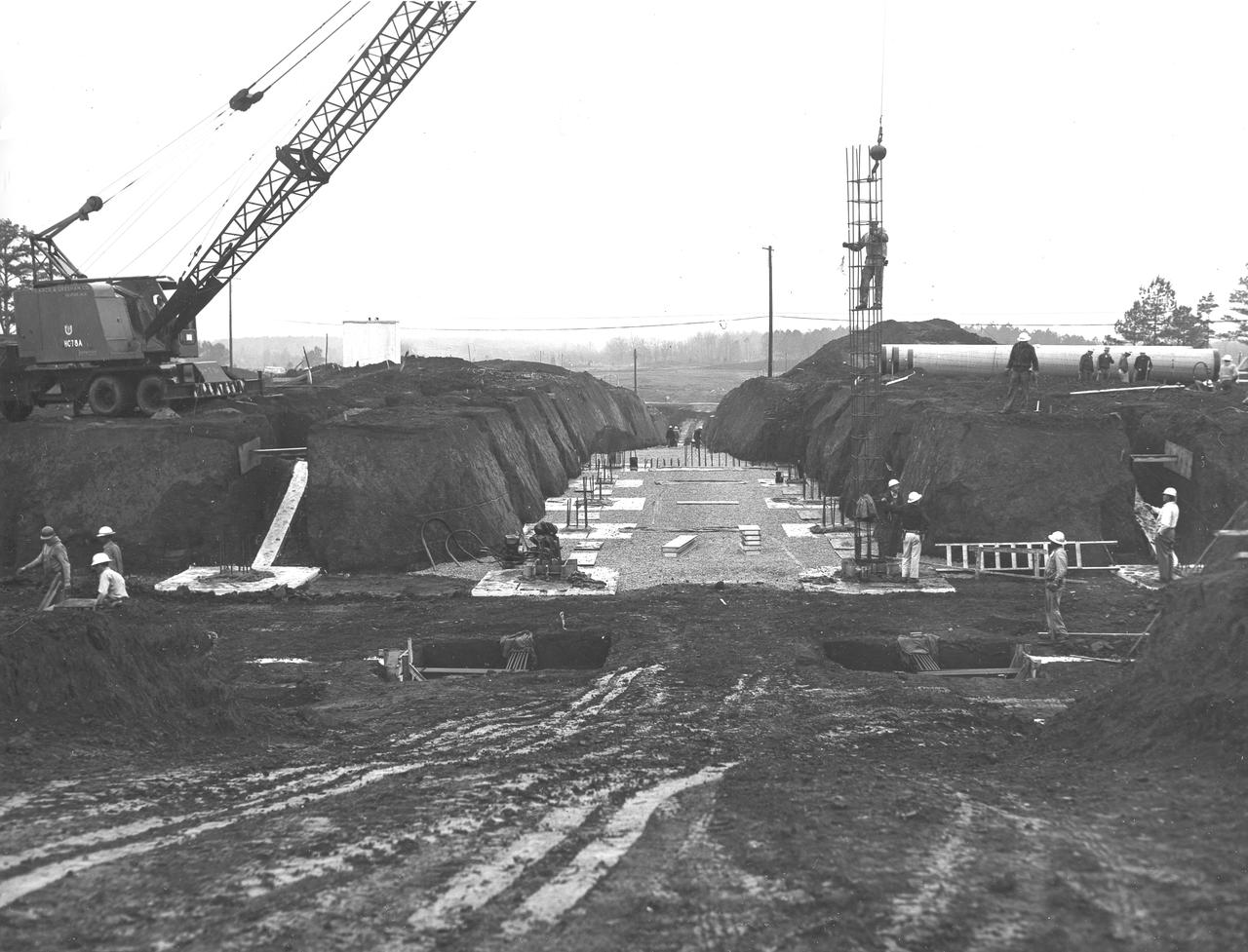 At its founding, the Marshall Space Flight Center (MSFC) inherited the Army’s Jupiter and Redstone test stands, but much larger facilities were needed for the giant stages of the Saturn V. From 1960 to 1964, the existing stands were remodeled and a sizable new test area was developed. The new comprehensive test complex for propulsion and structural dynamics was unique within the nation and the free world, and they remain so today because they were constructed with foresight to meet the future as well as on going needs. Construction of the S-IC Static test stand complex began in 1961 in the west test area of MSFC, and was completed in 1964. The S-IC static test stand was designed to develop and test the 138-ft long and 33-ft diameter Saturn V S-IC first stage, or booster stage, weighing in at 280,000 pounds. Required to hold down the brute force of a 7,500,000-pound thrust produced by 5 F-1 engines, the S-IC static test stand was designed and constructed with the strength of hundreds of tons of steel and 12,000,000 pounds of cement, planted down to bedrock 40 feet below ground level. The foundation walls, constructed with concrete and steel, are 4 feet thick. The base structure consists of four towers with 40-foot-thick walls extending upward 144 feet above ground level. The structure was topped by a crane with a 135-foot boom. With the boom in the upright position, the stand was given an overall height of 405 feet, placing it among the highest structures in Alabama at the time. In addition to the stand itself, related facilities were constructed during this time. Built directly east of the test stand was the Block House, which served as the control center for the test stand. The two were connected by a narrow tunnel which housed the cables for the controls. Again to the east, just south of the Block House, was a newly constructed Pump House. Its function was to provide water to the stand to prevent melting damage during testing. The water was sprayed through small holes in the stand’s 1900 ton water deflector at the rate of 320,000 gallons per minute. In this photo taken March 20, 1962, construction is well underway in the water line area connecting the Pump House to the S-IC test stand.