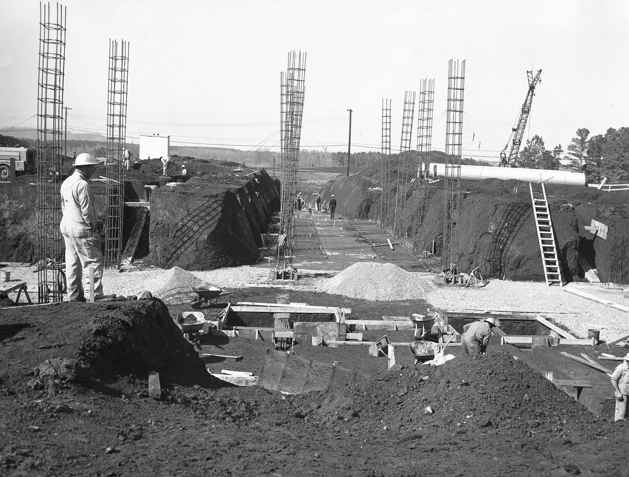 At its founding, the Marshall Space Flight Center (MSFC) inherited the Army’s Jupiter and Redstone test stands, but much larger facilities were needed for the giant stages of the Saturn V. From 1960 to 1964, the existing stands were remodeled and a sizable new test area was developed. The new comprehensive test complex for propulsion and structural dynamics was unique within the nation and the free world, and they remain so today because they were constructed with foresight to meet the future as well as on going needs. Construction of the S-IC Static test stand complex began in 1961 in the west test area of MSFC, and was completed in 1964. The S-IC static test stand was designed to develop and test the 138-ft long and 33-ft diameter Saturn V S-IC first stage, or booster stage, weighing in at 280,000 pounds. Required to hold down the brute force of a 7,500,000-pound thrust produced by 5 F-1 engines, the S-IC static test stand was designed and constructed with the strength of hundreds of tons of steel and 12,000,000 pounds of cement, planted down to bedrock 40 feet below ground level. The foundation walls, constructed with concrete and steel, are 4 feet thick. The base structure consists of four towers with 40-foot-thick walls extending upward 144 feet above ground level. The structure was topped by a crane with a 135-foot boom. With the boom in the upright position, the stand was given an overall height of 405 feet, placing it among the highest structures in Alabama at the time. In addition to the stand itself, related facilities were constructed during this time. Built directly east of the test stand was the Block House, which served as the control center for the test stand. The two were connected by a narrow tunnel which housed the cables for the controls. Again to the east, just south of the Block House, was a newly constructed Pump House. Its function was to provide water to the stand to prevent melting damage during testing. The water was sprayed through small holes in the stand’s 1900 ton water deflector at the rate of 320,000 gallons per minute. In this photo, taken March 20, 1962, construction of the Pump House area is well underway.  