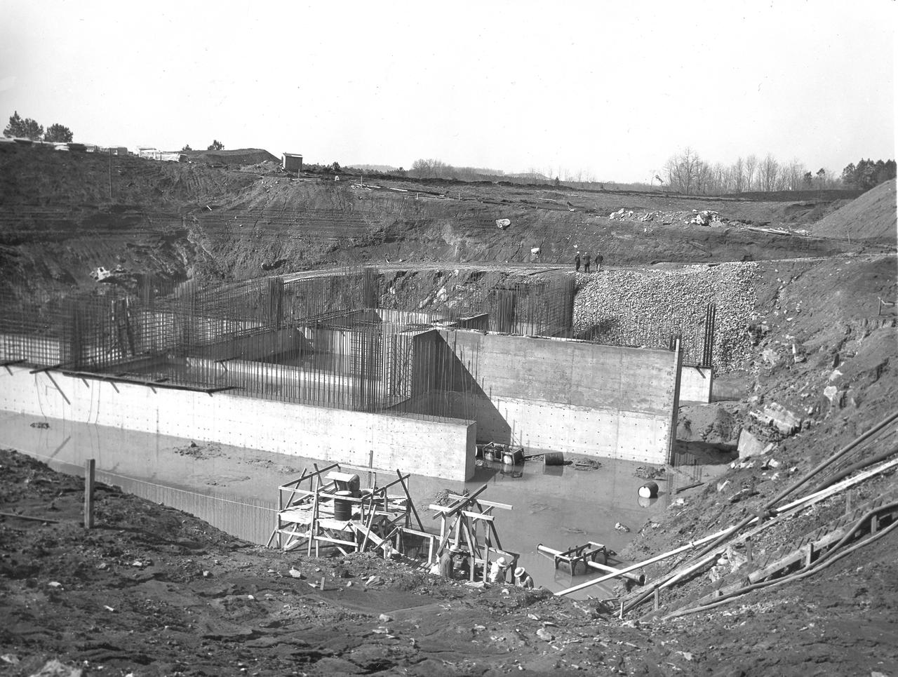 At its founding, the Marshall Space Flight Center (MSFC) inherited the Army’s Jupiter and Redstone test stands, but much larger facilities were needed for the giant stages of the Saturn V. From 1960 to 1964, the existing stands were remodeled and a sizable new test area was developed. The new comprehensive test complex for propulsion and structural dynamics was unique within the nation and the free world, and they remain so today because they were constructed with foresight to meet the future as well as on going needs. Construction of the S-IC Static test stand complex began in 1961 in the west test area of MSFC, and was completed in 1964. The S-IC static test stand was designed to develop and test the 138-ft long and 33-ft diameter Saturn V S-IC first stage, or booster stage, weighing in at 280,000 pounds. Required to hold down the brute force of a 7,500,000-pound thrust produced by 5 F-1 engines, the S-IC static test stand was designed and constructed with the strength of hundreds of tons of steel and 12,000,000 pounds of cement, planted down to bedrock 40 feet below ground level. The foundation walls, constructed with concrete and steel, are 4 feet thick. The base structure consists of four towers with 40-foot-thick walls extending upward 144 feet above ground level. The structure was topped by a crane with a 135-foot boom. With the boom in the upright position, the stand was given an overall height of 405 feet, placing it among the highest structures in Alabama at the time. Construction of the S-IC test stand came to a halt at the end of September 1961 as the determination was made that the Saturn booster size had to be increased. As a result, the stand had to be modified. With construction about to resume, portable, floating pump stations were placed in the site to drain the flood waters caused by a disturbed natural spring months prior during excavation. In this March 31, 1962 photo, the foundation walls can once again be seen.