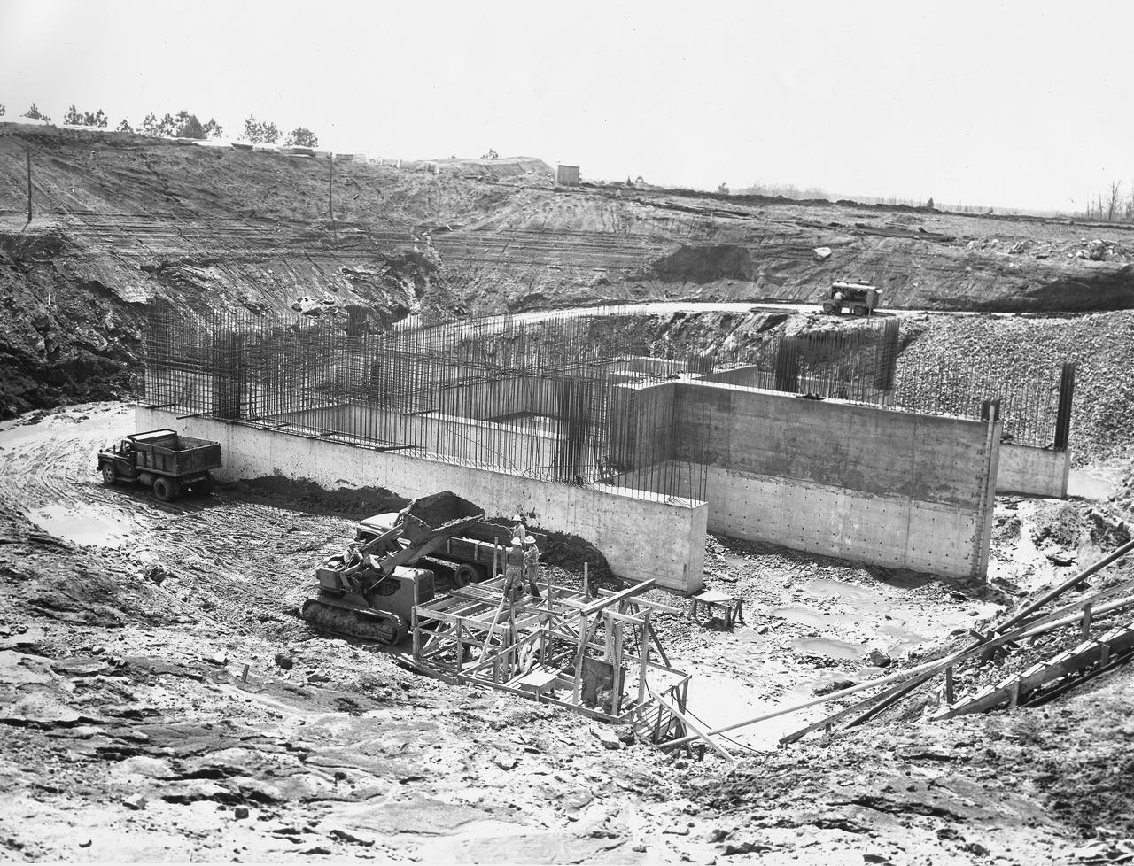 At its founding, the Marshall Space Flight Center (MSFC) inherited the Army’s Jupiter and Redstone test stands, but much larger facilities were needed for the giant stages of the Saturn V. From 1960 to 1964, the existing stands were remodeled and a sizable new test area was developed. The new comprehensive test complex for propulsion and structural dynamics was unique within the nation and the free world, and they remain so today because they were constructed with foresight to meet the future as well as on going needs. Construction of the S-IC Static test stand complex began in 1961 in the west test area of MSFC, and was completed in 1964. The S-IC static test stand was designed to develop and test the 138-ft long and 33-ft diameter Saturn V S-IC first stage, or booster stage, weighing in at 280,000 pounds. Required to hold down the brute force of a 7,500,000-pound thrust produced by 5 F-1 engines, the S-IC static test stand was designed and constructed with the strength of hundreds of tons of steel and 12,000,000 pounds of cement, planted down to bedrock 40 feet below ground level. The foundation walls, constructed with concrete and steel, are 4 feet thick. The base structure consists of four towers with 40-foot-thick walls extending upward 144 feet above ground level. The structure was topped by a crane with a 135-foot boom. With the boom in the upright position, the stand was given an overall height of 405 feet, placing it among the highest structures in Alabama at the time. This photo, taken April 4, 1961, shows the S-IC test stand dry once again when workers resumed construction after a 6 month delay due to booster size  reconfiguration back in September of 1961. The disturbance of a natural spring during the excavation of the site required water to be pumped from the site continuously. The site was completely flooded after the pumps were shut down during the construction delay.
