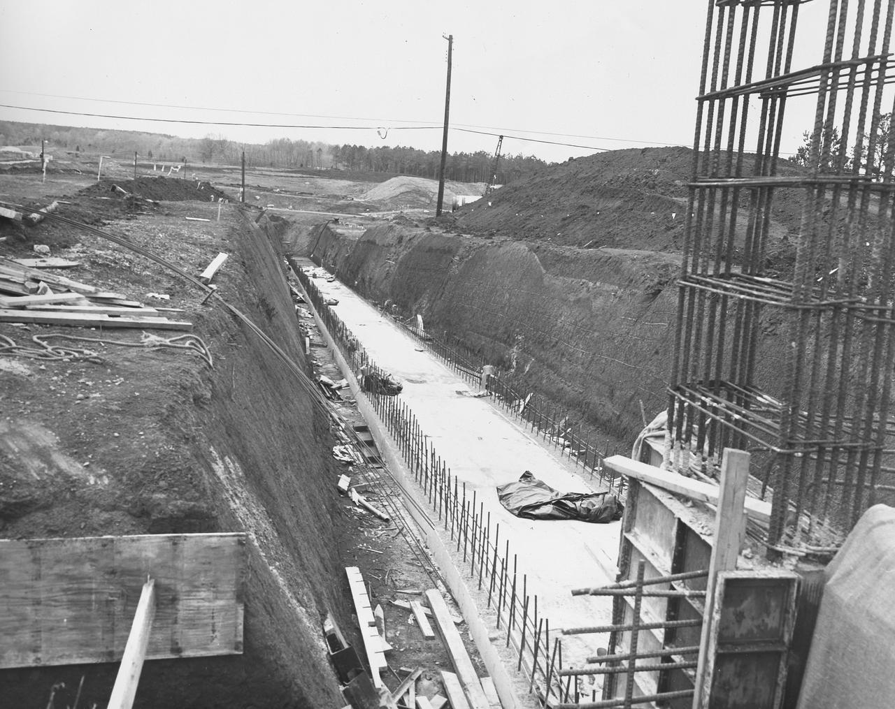 At its founding, the Marshall Space Flight Center (MSFC) inherited the Army’s Jupiter and Redstone test stands, but much larger facilities were needed for the giant stages of the Saturn V. From 1960 to 1964, the existing stands were remodeled and a sizable new test area was developed. The new comprehensive test complex for propulsion and structural dynamics was unique within the nation and the free world, and they remain so today because they were constructed with foresight to meet the future as well as on going needs. Construction of the S-IC Static test stand complex began in 1961 in the west test area of MSFC, and was completed in 1964. The S-IC static test stand was designed to develop and test the 138-ft long and 33-ft diameter Saturn V S-IC first stage, or booster stage, weighing in at 280,000 pounds. Required to hold down the brute force of a 7,500,000-pound thrust produced by 5 F-1 engines, the S-IC static test stand was designed and constructed with the strength of hundreds of tons of steel and 12,000,000 pounds of cement, planted down to bedrock 40 feet below ground level. The foundation walls, constructed with concrete and steel, are 4 feet thick. The base structure consists of four towers with 40-foot-thick walls extending upward 144 feet above ground level. The structure was topped by a crane with a 135-foot boom. With the boom in the upright position, the stand was given an overall height of 405 feet, placing it among the highest structures in Alabama at the time. In addition to the stand itself, related facilities were constructed during this time. Built directly east of the test stand was the Block House, which served as the control center for the test stand. The two were connected by a narrow tunnel which housed the cables for the controls. Again to the east, just south of the Block House, was a newly constructed Pump House. Its function was to provide water to the stand to prevent melting damage during testing. The water was sprayed through small holes in the stand’s 1900 ton water deflector at the rate of 320,000 gallons per minute. This photograph, taken Aril 16, 1962, depicts the water line area through which the pump house delivered water to the stand.
