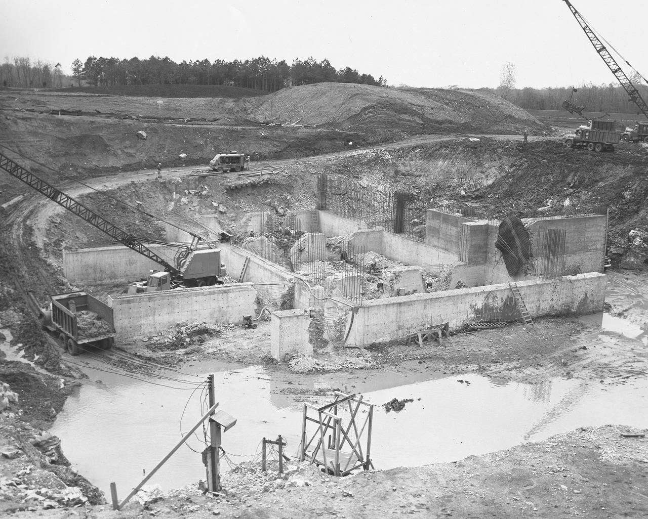 At its founding, the Marshall Space Flight Center (MSFC) inherited the Army’s Jupiter and Redstone test stands, but much larger facilities were needed for the giant stages of the Saturn V. From 1960 to 1964, the existing stands were remodeled and a sizable new test area was developed. The new comprehensive test complex for propulsion and structural dynamics was unique within the nation and the free world, and they remain so today because they were constructed with foresight to meet the future as well as on going needs. Construction of the S-IC Static test stand complex began in 1961 in the west test area of MSFC, and was completed in 1964. The S-IC static test stand was designed to develop and test the 138-ft long and 33-ft diameter Saturn V S-IC first stage, or booster stage, weighing in at 280,000 pounds. Required to hold down the brute force of a 7,500,000-pound thrust produced by 5 F-1 engines, the S-IC static test stand was designed and constructed with the strength of hundreds of tons of steel and 12,000,000 pounds of cement, planted down to bedrock 40 feet below ground level. The foundation walls, constructed with concrete and steel, are 4 feet thick. The base structure consists of four towers with 40-foot-thick walls extending upward 144 feet above ground level. The structure was topped by a crane with a 135-foot boom. With the boom in the upright position, the stand was given an overall height of 405 feet, placing it among the highest structures in Alabama at the time. After a 6 month delay in construction due to size reconfiguration of the Saturn booster, the site was revisited for modifications. The original foundation walls built in the prior year had to be torn down and re-poured to accommodate the larger booster. The demolition can be seen in this photograph taken on April 16, 1962.