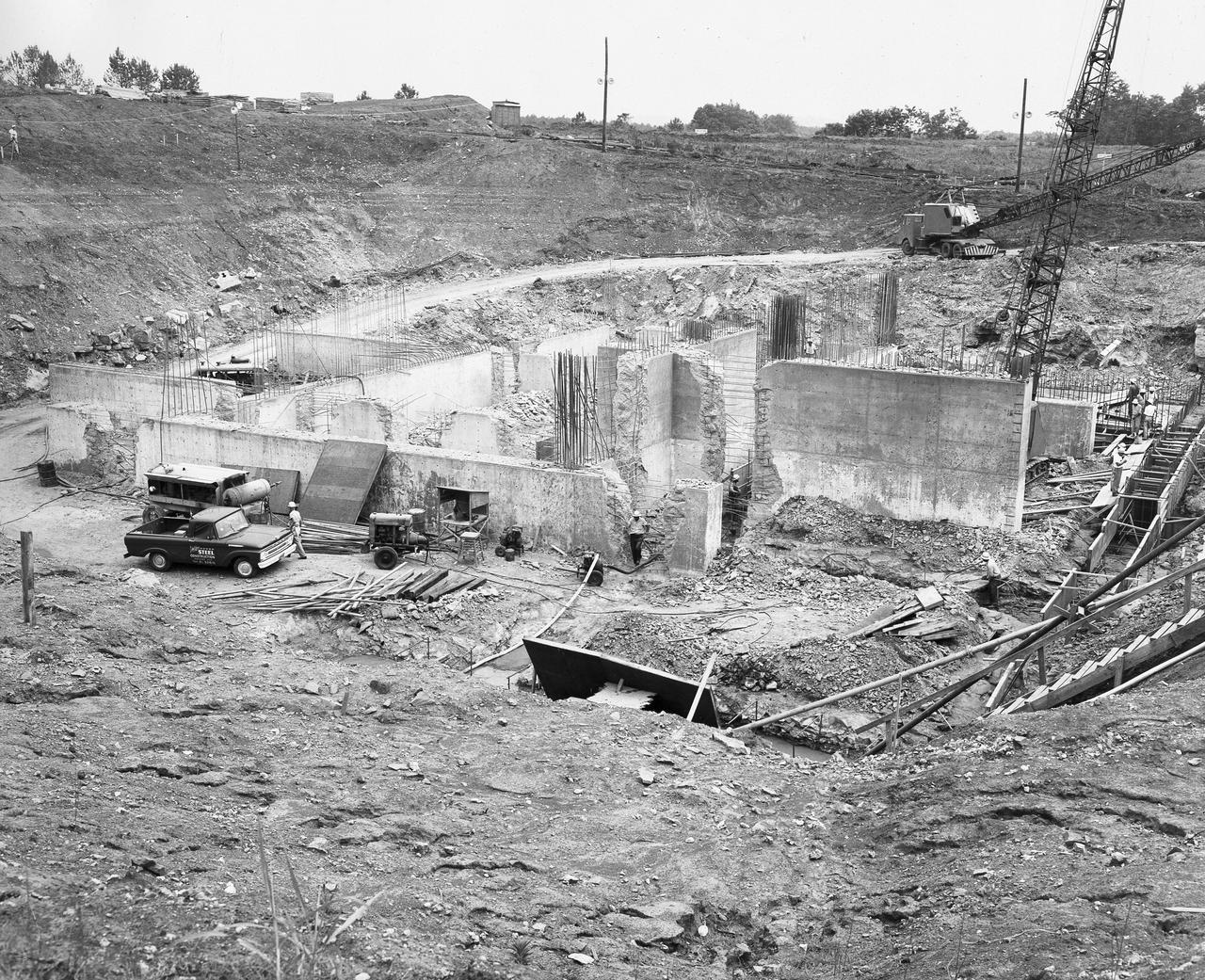 At its founding, the Marshall Space Flight Center (MSFC) inherited the Army’s Jupiter and Redstone test stands, but much larger facilities were needed for the giant stages of the Saturn V. From 1960 to 1964, the existing stands were remodeled and a sizable new test area was developed. The new comprehensive test complex for propulsion and structural dynamics was unique within the nation and the free world, and they remain so today because they were constructed with foresight to meet the future as well as on going needs. Construction of the S-IC Static test stand complex began in 1961 in the west test area of MSFC, and was completed in 1964. The S-IC static test stand was designed to develop and test the 138-ft long and 33-ft diameter Saturn V S-IC first stage, or booster stage, weighing in at 280,000 pounds. Required to hold down the brute force of a 7,500,000-pound thrust produced by 5 F-1 engines, the S-IC static test stand was designed and constructed with the strength of hundreds of tons of steel and 12,000,000 pounds of cement, planted down to bedrock 40 feet below ground level. The foundation walls, constructed with concrete and steel, are 4 feet thick. The base structure consists of four towers with 40-foot-thick walls extending upward 144 feet above ground level. The structure was topped by a crane with a 135-foot boom. With the boom in the upright position, the stand was given an overall height of 405 feet, placing it among the highest structures in Alabama at the time. After a 6 month delay in construction due to size reconfiguration of the Saturn booster, the site was revisited for modifications. The original foundation walls built in the prior year had to be torn down and re-poured to accommodate the larger booster. The demolition can be seen in this photograph taken on May 21, 1962.