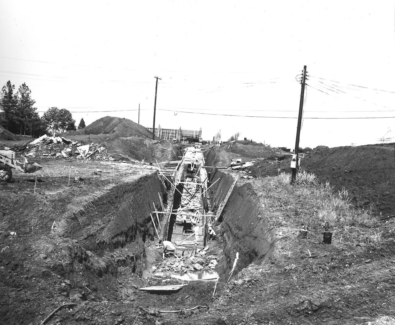 At its founding, the Marshall Space Flight Center (MSFC) inherited the Army’s Jupiter and Redstone test stands, but much larger facilities were needed for the giant stages of the Saturn V. From 1960 to 1964, the existing stands were remodeled and a sizable new test area was developed. The new comprehensive test complex for propulsion and structural dynamics was unique within the nation and the free world, and they remain so today because they were constructed with foresight to meet the future as well as on going needs. Construction of the S-IC Static test stand complex began in 1961 in the west test area of MSFC, and was completed in 1964. The S-IC static test stand was designed to develop and test the 138-ft long and 33-ft diameter Saturn V S-IC first stage, or booster stage, weighing in at 280,000 pounds. Required to hold down the brute force of a 7,500,000-pound thrust produced by 5 F-1 engines, the S-IC static test stand was designed and constructed with the strength of hundreds of tons of steel and 12,000,000 pounds of cement, planted down to bedrock 40 feet below ground level. The foundation walls, constructed with concrete and steel, are 4 feet thick. The base structure consists of four towers with 40-foot-thick walls extending upward 144 feet above ground level. The structure was topped by a crane with a 135-foot boom. With the boom in the upright position, the stand was given an overall height of 405 feet, placing it among the highest structures in Alabama at the time. In addition to the stand itself, related facilities were constructed during this time. Built directly east of the test stand was the Block House, which served as the control center for the test stand. The two were connected by a narrow access tunnel which housed the cables for the controls. Construction of the tunnel is depicted in this photo taken June 13, 1962.