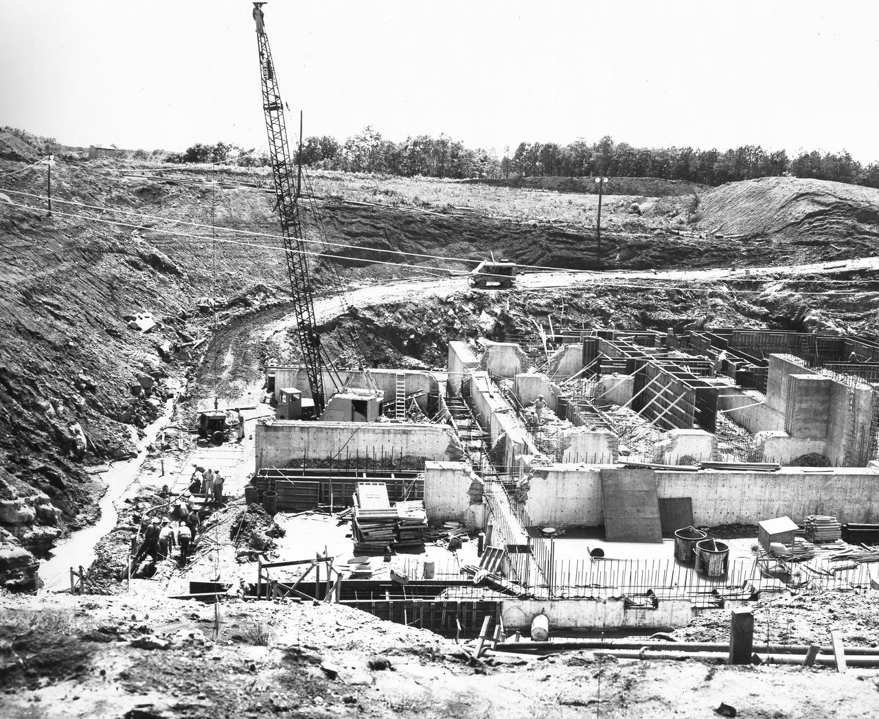 At its founding, the Marshall Space Flight Center (MSFC) inherited the Army’s Jupiter and Redstone test stands, but much larger facilities were needed for the giant stages of the Saturn V. From 1960 to 1964, the existing stands were remodeled and a sizable new test area was developed. The new comprehensive test complex for propulsion and structural dynamics was unique within the nation and the free world, and they remain so today because they were constructed with foresight to meet the future as well as on going needs. Construction of the S-IC Static test stand complex began in 1961 in the west test area of MSFC, and was completed in 1964. The S-IC static test stand was designed to develop and test the 138-ft long and 33-ft diameter Saturn V S-IC first stage, or booster stage, weighing in at 280,000 pounds. Required to hold down the brute force of a 7,500,000-pound thrust produced by 5 F-1 engines, the S-IC static test stand was designed and constructed with the strength of hundreds of tons of steel and 12,000,000 pounds of cement, planted down to bedrock 40 feet below ground level. The foundation walls, constructed with concrete and steel, are 4 feet thick. The base structure consists of four towers with 40-foot-thick walls extending upward 144 feet above ground level. The structure was topped by a crane with a 135-foot boom. With the boom in the upright position, the stand was given an overall height of 405 feet, placing it among the highest structures in Alabama at the time. After a six month delay in construction due to size reconfiguration of the Saturn booster, the site was revisited for modifications in March 1962. The original foundation walls built in the prior year were torn down and re-poured to accommodate the larger boosters. This photo depicts that modification progress as of June 13,1962. 