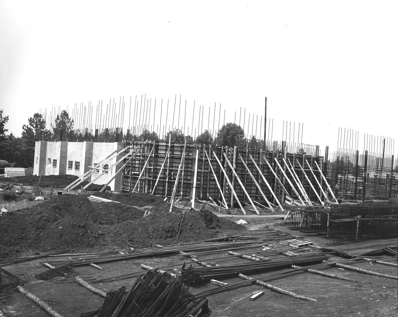 At its founding, the Marshall Space Flight Center (MSFC) inherited the Army’s Jupiter and Redstone test stands, but much larger facilities were needed for the giant stages of the Saturn V. From 1960 to 1964, the existing stands were remodeled and a sizable new test area was developed. The new comprehensive test complex for propulsion and structural dynamics was unique within the nation and the free world, and they remain so today because they were constructed with foresight to meet the future as well as on going needs. Construction of the S-IC Static test stand complex began in 1961 in the west test area of MSFC, and was completed in 1964. The S-IC static test stand was designed to develop and test the 138-ft long and 33-ft diameter Saturn V S-IC first stage, or booster stage, weighing in at 280,000 pounds. Required to hold down the brute force of a 7,500,000-pound thrust produced by 5 F-1 engines, the S-IC static test stand was designed and constructed with the strength of hundreds of tons of steel and 12,000,000 pounds of cement, planted down to bedrock 40 feet below ground level. The foundation walls, constructed with concrete and steel, are 4 feet thick. The base structure consists of four towers with 40-foot-thick walls extending upward 144 feet above ground level. The structure was topped by a crane with a 135-foot boom. With the boom in the upright position, the stand was given an overall height of 405 feet, placing it among the highest structures in Alabama at the time. In addition to the stand itself, related facilities were constructed during this time. Built directly east of the test stand was the Block House, which served as the control center for the test stand. The two were connected by a narrow access tunnel which housed the cables for the controls. This construction photo taken July 3, 1962 depicts the Block House with a portion of its concrete walls poured and exposed while many are still in the forms stage.