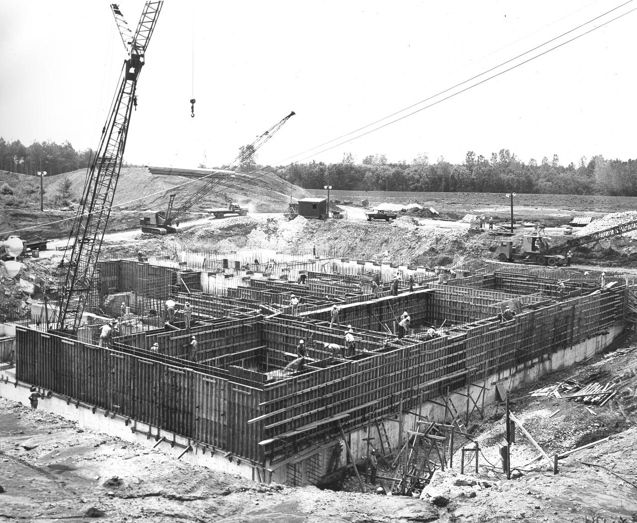 At its founding, the Marshall Space Flight Center (MSFC) inherited the Army’s Jupiter and Redstone test stands, but much larger facilities were needed for the giant stages of the Saturn V. From 1960 to 1964, the existing stands were remodeled and a sizable new test area was developed. The new comprehensive test complex for propulsion and structural dynamics was unique within the nation and the free world, and they remain so today because they were constructed with foresight to meet the future as well as on going needs. Construction of the S-IC Static test stand complex began in 1961 in the west test area of MSFC, and was completed in 1964. The S-IC static test stand was designed to develop and test the 138-ft long and 33-ft diameter Saturn V S-IC first stage, or booster stage, weighing in at 280,000 pounds. Required to hold down the brute force of a 7,500,000-pound thrust produced by 5 F-1 engines, the S-IC static test stand was designed and constructed with the strength of hundreds of tons of steel and 12,000,000 pounds of cement, planted down to bedrock 40 feet below ground level. The foundation walls, constructed with concrete and steel, are 4 feet thick. The base structure consists of four towers with 40-foot-thick walls extending upward 144 feet above ground level. The structure was topped by a crane with a 135-foot boom. With the boom in the upright position, the stand was given an overall height of 405 feet, placing it among the highest structures in Alabama at the time.  In addition to the stand itself, related facilities were constructed during this time. Built directly east of the test stand was the Block House, which served as the control center for the test stand. The two were connected by a narrow access tunnel which housed the cables for the controls. This construction photo, taken July 3, 1962, depicts the Block House with a portion of its concrete walls poured and exposed while many are still in the forms stage.