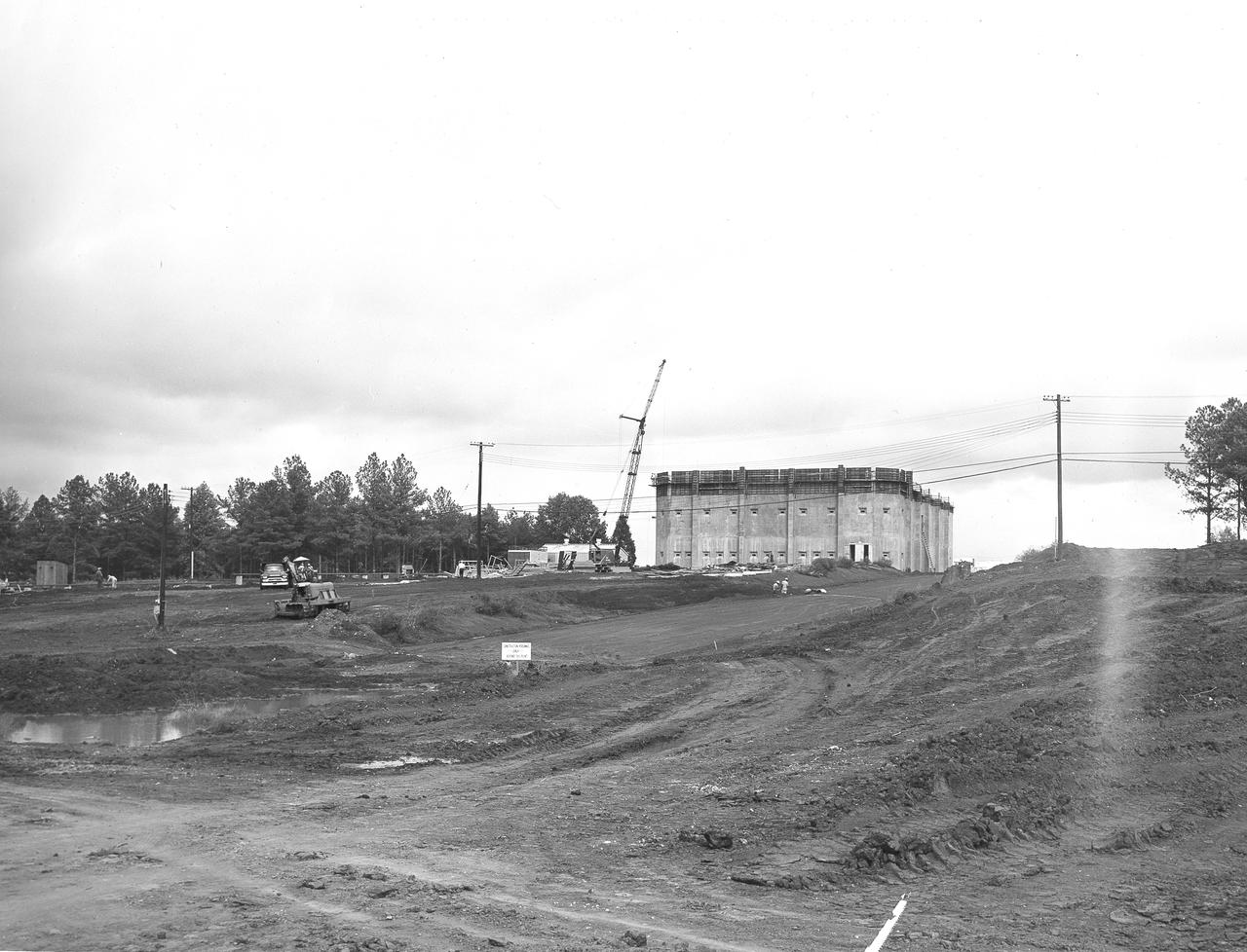 At its founding, the Marshall Space Flight Center (MSFC) inherited the Army’s Jupiter and Redstone test stands, but much larger facilities were needed for the giant stages of the Saturn V. From 1960 to 1964, the existing stands were remodeled and a sizable new test area was developed. The new comprehensive test complex for propulsion and structural dynamics was unique within the nation and the free world, and they remain so today because they were constructed with foresight to meet the future as well as on going needs. Construction of the S-IC Static test stand complex began in 1961 in the west test area of MSFC, and was completed in 1964. The S-IC static test stand was designed to develop and test the 138-ft long and 33-ft diameter Saturn V S-IC first stage, or booster stage, weighing in at 280,000 pounds. Required to hold down the brute force of a 7,500,000-pound thrust produced by 5 F-1 engines, the S-IC static test stand was designed and constructed with the strength of hundreds of tons of steel and 12,000,000 pounds of cement, planted down to bedrock 40 feet below ground level. The foundation walls, constructed with concrete and steel, are 4 feet thick. The base structure consists of four towers with 40-foot-thick walls extending upward 144 feet above ground level. The structure was topped by a crane with a 135-foot boom. With the boom in the upright position, the stand was given an overall height of 405 feet, placing it among the highest structures in Alabama at the time.  Built directly east of the test stand was the Block House, which served as the control center for the test stand. The two were connected by a narrow access tunnel which housed the cables for the controls. This construction photo, taken October 8, 1962, depicts a front view of the Block House nearing completion.