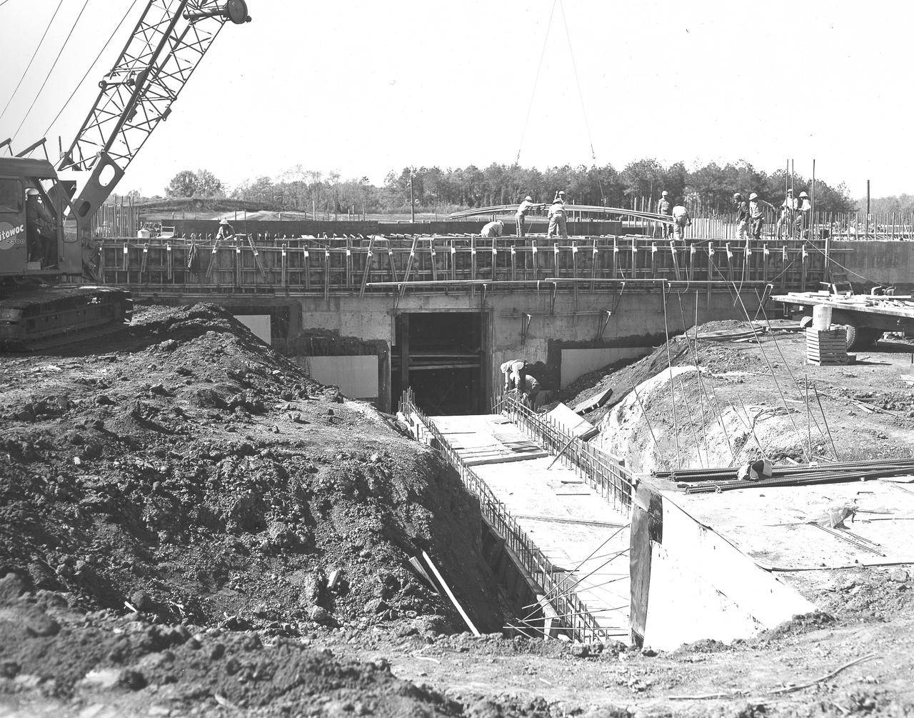 At its founding, the Marshall Space Flight Center (MSFC) inherited the Army’s Jupiter and Redstone test stands, but much larger facilities were needed for the giant stages of the Saturn V. From 1960 to 1964, the existing stands were remodeled and a sizable new test area was developed. The new comprehensive test complex for propulsion and structural dynamics was unique within the nation and the free world, and they remain so today because they were constructed with foresight to meet the future as well as on going needs. Construction of the S-IC Static test stand complex began in 1961 in the west test area of MSFC, and was completed in 1964. The S-IC static test stand was designed to develop and test the 138-ft long and 33-ft diameter Saturn V S-IC first stage, or booster stage, weighing in at 280,000 pounds. Required to hold down the brute force of a 7,500,000-pound thrust produced by 5 F-1 engines, the S-IC static test stand was designed and constructed with the strength of hundreds of tons of steel and 12,000,000 pounds of cement, planted down to bedrock 40 feet below ground level. The foundation walls, constructed with concrete and steel, are 4 feet thick. The base structure consists of four towers with 40-foot-thick walls extending upward 144 feet above ground level. The structure was topped by a crane with a 135-foot boom. With the boom in the upright position, the stand was given an overall height of 405 feet, placing it among the highest structures in Alabama at the time. In addition to the stand itself, related facilities were constructed during this time. Built directly east of the test stand was the Block House, which served as the control center for the test stand. The two were connected by a narrow access tunnel which housed the cables for the controls. This construction photo, taken October 26, 1962, depicts a view of the Block House tunnel opening.
