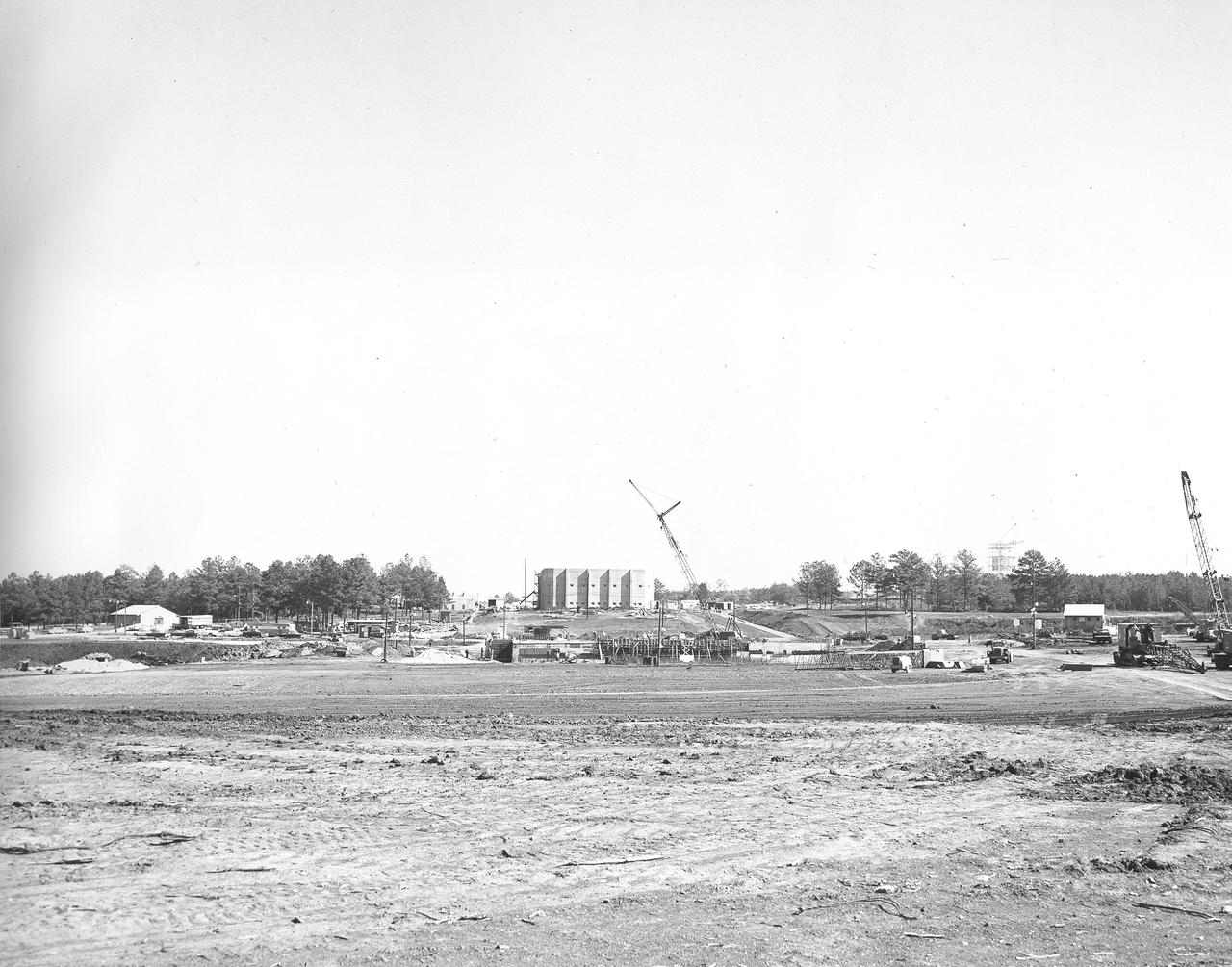 At its founding, the Marshall Space Flight Center (MSFC) inherited the Army’s Jupiter and Redstone test stands, but much larger facilities were needed for the giant stages of the Saturn V. From 1960 to 1964, the existing stands were remodeled and a sizable new test area was developed. The new comprehensive test complex for propulsion and structural dynamics was unique within the nation and the free world, and they remain so today because they were constructed with foresight to meet the future as well as on going needs. Construction of the S-IC Static test stand complex began in 1961 in the west test area of MSFC, and was completed in 1964. The S-IC static test stand was designed to develop and test the 138-ft long and 33-ft diameter Saturn V S-IC first stage, or booster stage, weighing in at 280,000 pounds. Required to hold down the brute force of a 7,500,000-pound thrust produced by 5 F-1 engines, the S-IC static test stand was designed and constructed with the strength of hundreds of tons of steel and 12,000,000 pounds of cement, planted down to bedrock 40 feet below ground level. The foundation walls, constructed with concrete and steel, are 4 feet thick. The base structure consists of four towers with 40-foot-thick walls extending upward 144 feet above ground level. The structure was topped by a crane with a 135-foot boom. With the boom in the upright position, the stand was given an overall height of 405 feet, placing it among the highest structures in Alabama at the time. In addition to the stand itself, related facilities were constructed during this time. Built directly east of the test stand was the Block House, which served as the control center for the test stand. The two were connected by a narrow access tunnel which housed the cables for the controls. This distant construction photo, taken October 26, 1962, depicts a view of the Block House and test stand site.