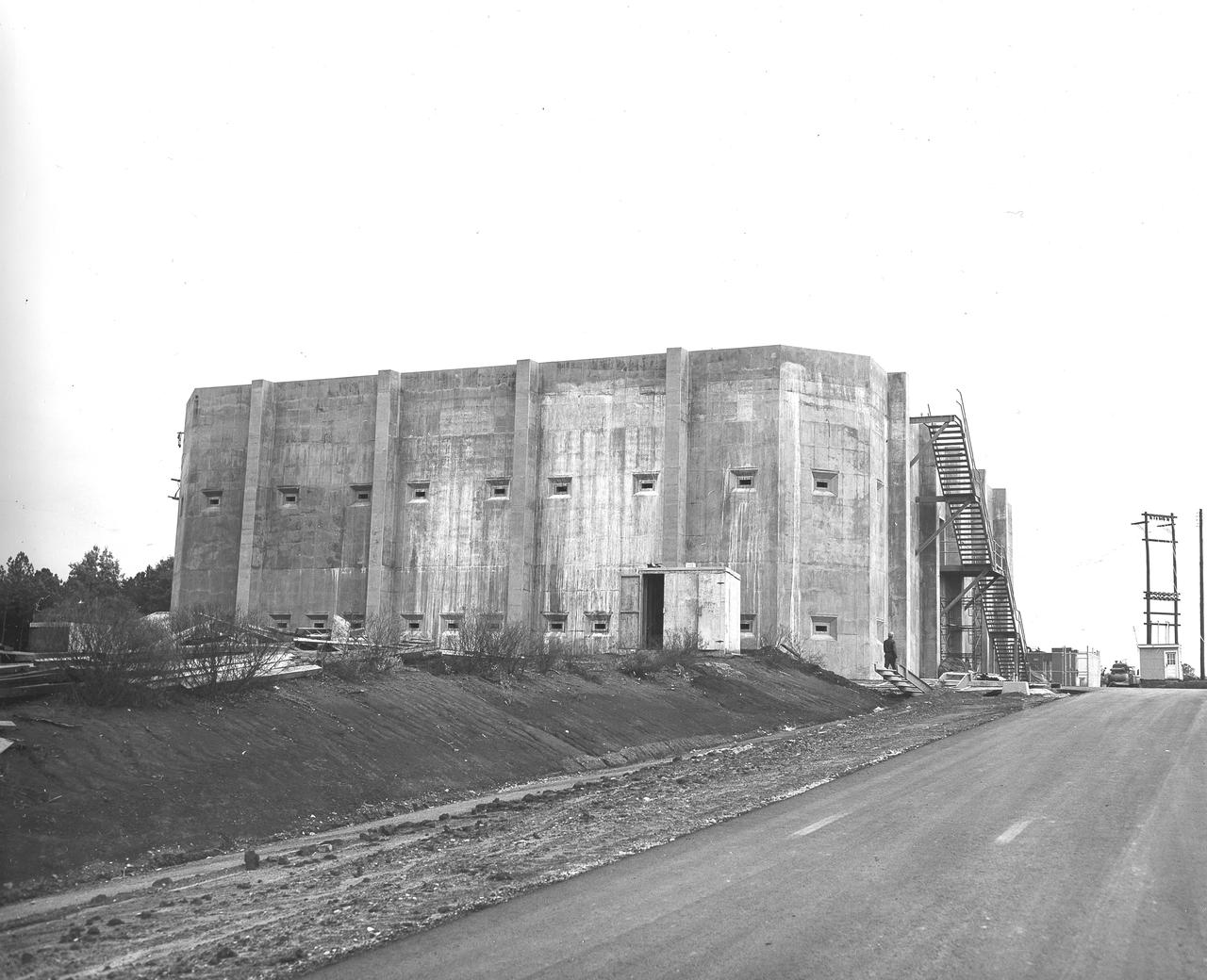 At its founding, the Marshall Space Flight Center (MSFC) inherited the Army’s Jupiter and Redstone test stands, but much larger facilities were needed for the giant stages of the Saturn V. From 1960 to 1964, the existing stands were remodeled and a sizable new test area was developed. The new comprehensive test complex for propulsion and structural dynamics was unique within the nation and the free world, and they remain so today because they were constructed with foresight to meet the future as well as on going needs. Construction of the S-IC Static test stand complex began in 1961 in the west test area of MSFC, and was completed in 1964. The S-IC static test stand was designed to develop and test the 138-ft long and 33-ft diameter Saturn V S-IC first stage, or booster stage, weighing in at 280,000 pounds. Required to hold down the brute force of a 7,500,000-pound thrust produced by 5 F-1 engines, the S-IC static test stand was designed and constructed with the strength of hundreds of tons of steel and 12,000,000 pounds of cement, planted down to bedrock 40 feet below ground level. The foundation walls, constructed with concrete and steel, are 4 feet thick. The base structure consists of four towers with 40-foot-thick walls extending upward 144 feet above ground level. The structure was topped by a crane with a 135-foot boom. With the boom in the upright position, the stand was given an overall height of 405 feet, placing it among the highest structures in Alabama at the time. In addition to the stand itself, related facilities were constructed during this time. Built directly east of the test stand was the Block House, which served as the control center for the test stand. The two were connected by a narrow access tunnel which housed the cables for the controls. This construction photo, taken November 15, 1962, depicts a view of the Block House.