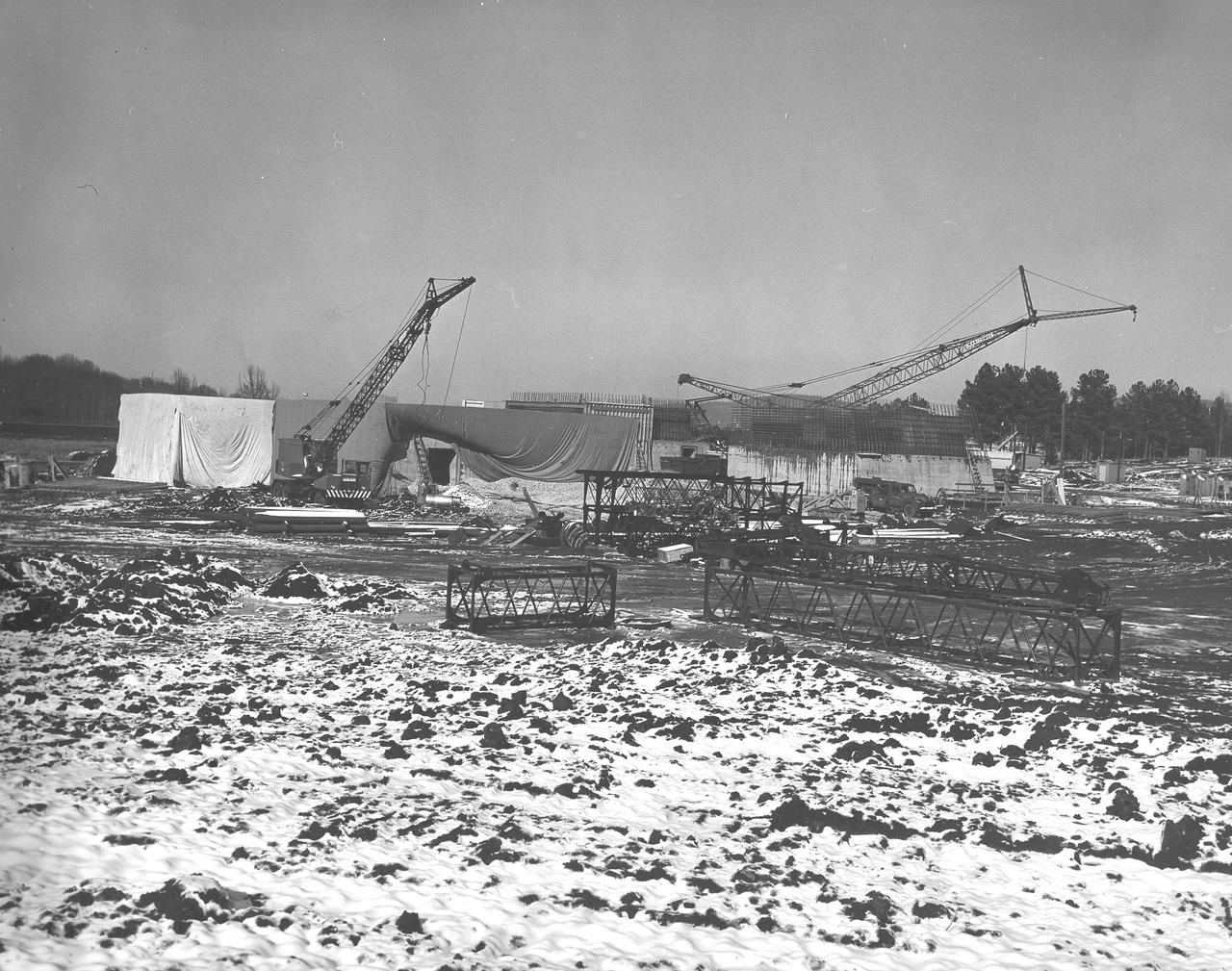 At its founding, the Marshall Space Flight Center (MSFC) inherited the Army’s Jupiter and Redstone test stands, but much larger facilities were needed for the giant stages of the Saturn V. From 1960 to 1964, the existing stands were remodeled and a sizable new test area was developed. The new comprehensive test complex for propulsion and structural dynamics was unique within the nation and the free world, and they remain so today because they were constructed with foresight to meet the future as well as on going needs. Construction of the S-IC Static test stand complex began in 1961 in the west test area of MSFC, and was completed in 1964. The S-IC static test stand was designed to develop and test the 138-ft long and 33-ft diameter Saturn V S-IC first stage, or booster stage, weighing in at 280,000 pounds. Required to hold down the brute force of a 7,500,000-pound thrust produced by 5 F-1 engines, the S-IC static test stand was designed and constructed with the strength of hundreds of tons of steel and 12,000,000 pounds of cement, planted down to bedrock 40 feet below ground level. The foundation walls, constructed with concrete and steel, are 4 feet thick. The base structure consists of four towers with 40-foot-thick walls extending upward 144 feet above ground level. The structure was topped by a crane with a 135-foot boom. With the boom in the upright position, the stand was given an overall height of 405 feet, placing it among the highest structures in Alabama at the time. This photo, depicts the progress of the stand as of January 14, 1963, with its four towers prominently rising.