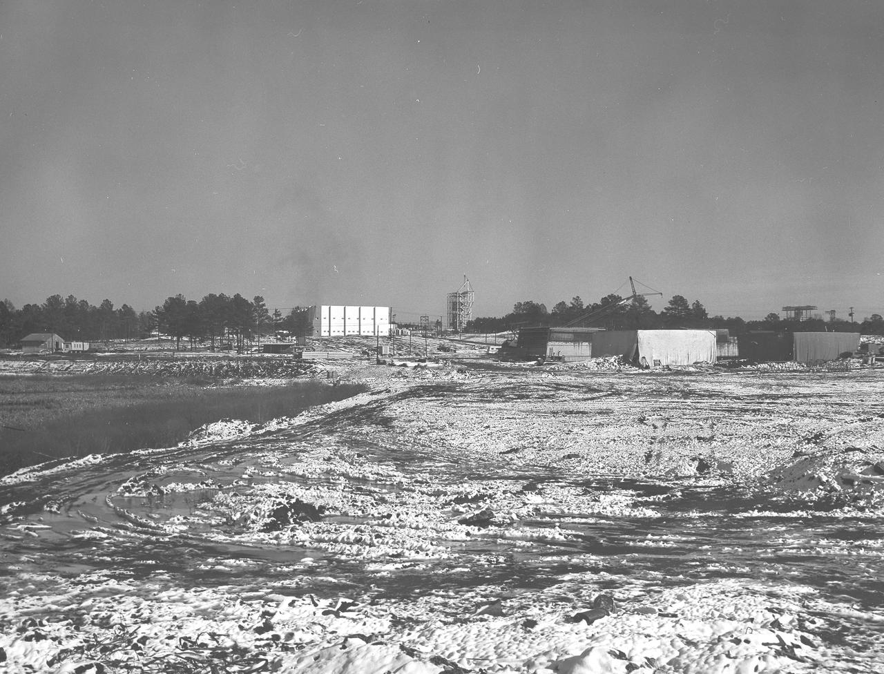 At its founding, the Marshall Space Flight Center (MSFC) inherited the Army’s Jupiter and Redstone test stands, but much larger facilities were needed for the giant stages of the Saturn V. From 1960 to 1964, the existing stands were remodeled and a sizable new test area was developed. The new comprehensive test complex for propulsion and structural dynamics was unique within the nation and the free world, and they remain so today because they were constructed with foresight to meet the future as well as on going needs. Construction of the S-IC Static test stand complex began in 1961 in the west test area of MSFC, and was completed in 1964. The S-IC static test stand was designed to develop and test the 138-ft long and 33-ft diameter Saturn V S-IC first stage, or booster stage, weighing in at 280,000 pounds. Required to hold down the brute force of a 7,500,000-pound thrust produced by 5 F-1 engines, the S-IC static test stand was designed and constructed with the strength of hundreds of tons of steel and 12,000,000 pounds of cement, planted down to bedrock 40 feet below ground level. The foundation walls, constructed with concrete and steel, are 4 feet thick. The base structure consists of four towers with 40-foot-thick walls extending upward 144 feet above ground level. The structure was topped by a crane with a 135-foot boom. With the boom in the upright position, the stand was given an overall height of 405 feet, placing it among the highest structures in Alabama at the time. This photo, depicts the progress of the stand as of January 14, 1963, with its four towers prominently rising.