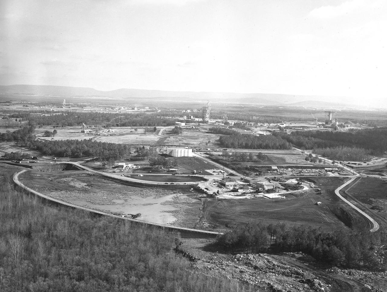 At its founding, the Marshall Space Flight Center (MSFC) inherited the Army’s Jupiter and Redstone test stands, but much larger facilities were needed for the giant stages of the Saturn V. From 1960 to 1964, the existing stands were remodeled and a sizable new test area was developed. The new comprehensive test complex for propulsion and structural dynamics was unique within the nation and the free world, and they remain so today because they were constructed with foresight to meet the future as well as on going needs. Construction of the S-IC Static test stand complex began in 1961 in the west test area of MSFC, and was completed in 1964. The S-IC static test stand was designed to develop and test the 138-ft long and 33-ft diameter Saturn V S-IC first stage, or booster stage, weighing in at 280,000 pounds. Required to hold down the brute force of a 7,500,000-pound thrust produced by 5 F-1 engines, the S-IC static test stand was designed and constructed with the strength of hundreds of tons of steel and 12,000,000 pounds of cement, planted down to bedrock 40 feet below ground level. The foundation walls, constructed with concrete and steel, are 4 feet thick. The base structure consists of four towers with 40-foot-thick walls extending upward 144 feet above ground level. The structure was topped by a crane with a 135-foot boom. With the boom in the upright position, the stand was given an overall height of 405 feet, placing it among the highest structures in Alabama at the time. In addition to the stand itself, related facilities were constructed during this time. Built directly east of the test stand was the Block House, which served as the control center for the test stand. The two were connected by a narrow access tunnel which housed the cables for the controls. The F-1 Engine test stand was built north of the massive S-IC test stand. The F-1 test stand is a vertical engine firing test stand, 239 feet in elevation and 4,600 square feet in area at the base, and was designed to assist in the development of the F-1 Engine. Capability is provided for static firing of 1.5 million pounds of thrust using liquid oxygen and kerosene. Like the S-IC stand, the foundation of the F-1 stand is keyed into the bedrock approximately 40 feet below grade. This aerial photograph, taken January 15, 1963 gives an overall view of the construction progress of the newly developed test complex. The large white building located in the center is the Block House. Just below and to the right of it is the S-IC test stand. The large hole to the left of the S-IC stand is the F-1 test stand site.