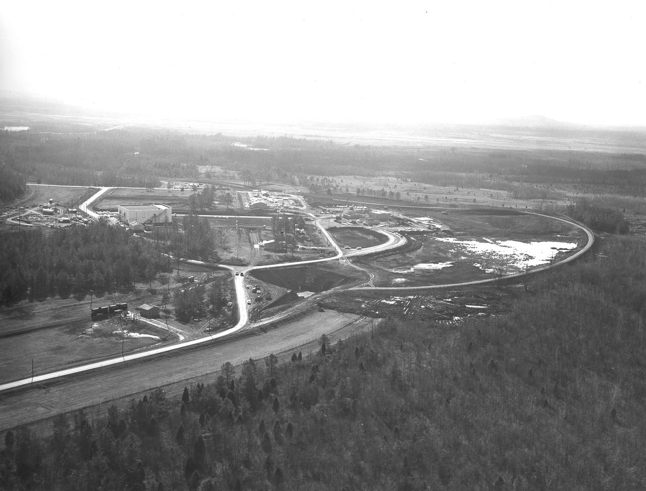 At its founding, the Marshall Space Flight Center (MSFC) inherited the Army’s Jupiter and Redstone test stands, but much larger facilities were needed for the giant stages of the Saturn V. From 1960 to 1964, the existing stands were remodeled and a sizable new test area was developed. The new comprehensive test complex for propulsion and structural dynamics was unique within the nation and the free world, and they remain so today because they were constructed with foresight to meet the future as well as on going needs. Construction of the S-IC Static test stand complex began in 1961 in the west test area of MSFC, and was completed in 1964. The S-IC static test stand was designed to develop and test the 138-ft long and 33-ft diameter Saturn V S-IC first stage, or booster stage, weighing in at 280,000 pounds. Required to hold down the brute force of a 7,500,000-pound thrust produced by 5 F-1 engines, the S-IC static test stand was designed and constructed with the strength of hundreds of tons of steel and 12,000,000 pounds of cement, planted down to bedrock 40 feet below ground level. The foundation walls, constructed with concrete and steel, are 4 feet thick. The base structure consists of four towers with 40-foot-thick walls extending upward 144 feet above ground level. The structure was topped by a crane with a 135-foot boom. With the boom in the upright position, the stand was given an overall height of 405 feet, placing it among the highest structures in Alabama at the time. In addition to the stand itself, related facilities were constructed during this time. Built directly east of the test stand was the Block House, which served as the control center for the test stand. The two were connected by a narrow access tunnel which housed the cables for the controls. The F-1 Engine test stand was built north of the massive S-IC test stand. The F-1 test stand is a vertical engine firing test stand, 239 feet in elevation and 4,600 square feet in area at the base, and was designed to assist in the development of the F-1 Engine. Capability is provided for static firing of 1.5 million pounds of thrust using liquid oxygen and kerosene. Like the S-IC stand, the foundation of the F-1 stand is keyed into the bedrock approximately 40 feet below grade. Looking North, this aerial taken January 15, 1963, gives a closer view of the deep hole for the F-1 test stand site in the forefront. The S-IC test stand with towers prominent is to the right of center, and the Block House is seen left of center.