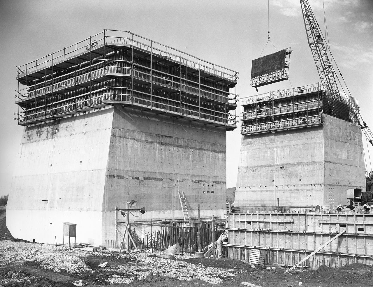 At its founding, the Marshall Space Flight Center (MSFC) inherited the Army’s Jupiter and Redstone test stands, but much larger facilities were needed for the giant stages of the Saturn V. From 1960 to 1964, the existing stands were remodeled and a sizable new test area was developed. The new comprehensive test complex for propulsion and structural dynamics was unique within the nation and the free world, and they remain so today because they were constructed with foresight to meet the future as well as on going needs. Construction of the S-IC Static test stand complex began in 1961 in the west test area of MSFC, and was completed in 1964. The S-IC static test stand was designed to develop and test the 138-ft long and 33-ft diameter Saturn V S-IC first stage, or booster stage, weighing in at 280,000 pounds. Required to hold down the brute force of a 7,500,000-pound thrust produced by 5 F-1 engines, the S-IC static test stand was designed and constructed with the strength of hundreds of tons of steel and 12,000,000 pounds of cement, planted down to bedrock 40 feet below ground level. The foundation walls, constructed with concrete and steel, are 4 feet thick. The base structure consists of four towers with 40-foot-thick walls extending upward 144 feet above ground level. The structure was topped by a crane with a 135-foot boom. With the boom in the upright position, the stand was given an overall height of 405 feet, placing it among the highest structures in Alabama at the time. This photograph taken February 25, 1963, gives a close up look at two of the ever-growing four towers of the S-IC Test Stand.