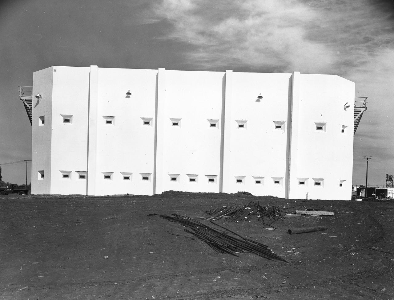 At its founding, the Marshall Space Flight Center (MSFC) inherited the Army’s Jupiter and Redstone test stands, but much larger facilities were needed for the giant stages of the Saturn V. From 1960 to 1964, the existing stands were remodeled and a sizable new test area was developed. The new comprehensive test complex for propulsion and structural dynamics was unique within the nation and the free world, and they remain so today because they were constructed with foresight to meet the future as well as on going needs. Construction of the S-IC Static test stand complex began in 1961 in the west test area of MSFC, and was completed in 1964. The S-IC static test stand was designed to develop and test the 138-ft long and 33-ft diameter Saturn V S-IC first stage, or booster stage, weighing in at 280,000 pounds. Required to hold down the brute force of a 7,500,000-pound thrust produced by 5 F-1 engines, the S-IC static test stand was designed and constructed with the strength of hundreds of tons of steel and 12,000,000 pounds of cement, planted down to bedrock 40 feet below ground level. The foundation walls, constructed with concrete and steel, are 4 feet thick. The base structure consists of four towers with 40-foot-thick walls extending upward 144 feet above ground level. The structure was topped by a crane with a 135-foot boom. With the boom in the upright position, the stand was given an overall height of 405 feet, placing it among the highest structures in Alabama at the time. In addition to the stand itself, related facilities were constructed during this time. Built directly east of the test stand was the Block House, which served as the control center for the test stand. The two were connected by a narrow access tunnel which housed the cables for the controls. This photograph, taken February 25, 1963, gives a close up look at the completed Block House. The side shown faces the S-IC Test Stand.