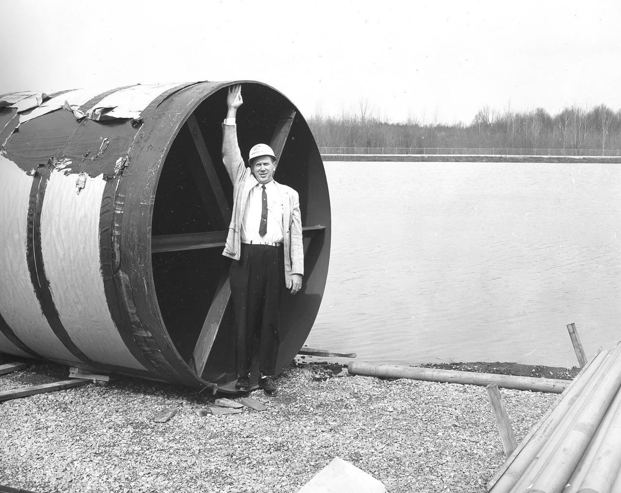At its founding, the Marshall Space Flight Center (MSFC) inherited the Army’s Jupiter and Redstone test stands, but much larger facilities were needed for the giant stages of the Saturn V. From 1960 to 1964, the existing stands were remodeled and a sizable new test area was developed. The new comprehensive test complex for propulsion and structural dynamics was unique within the nation and the free world, and they remain so today because they were constructed with foresight to meet the future as well as on going needs. Construction of the S-IC Static test stand complex began in 1961 in the west test area of MSFC, and was completed in 1964. The S-IC static test stand was designed to develop and test the 138-ft long and 33-ft diameter Saturn V S-IC first stage, or booster stage, weighing in at 280,000 pounds. Required to hold down the brute force of a 7,500,000-pound thrust produced by 5 F-1 engines, the S-IC static test stand was designed and constructed with the strength of hundreds of tons of steel and 12,000,000 pounds of cement, planted down to bedrock 40 feet below ground level. The foundation walls, constructed with concrete and steel, are 4 feet thick. The base structure consists of four towers with 40-foot-thick walls extending upward 144 feet above ground level. The structure was topped by a crane with a 135-foot boom. With the boom in the upright position, the stand was given an overall height of 405 feet, placing it among the highest structures in Alabama at the time. In addition to the stand itself, related facilities were constructed during this time. Built directly east of the test stand was the Block House, which served as the control center for the test stand. The two were connected by a narrow access tunnel which housed the cables for the controls. Again to the east, just south of the Block House, was a newly constructed Pump House. Its function was to provide water to the stand to prevent melting damage during testing. The water was sprayed through small holes in the stand’s 1900 ton water deflector at the rate of 320,000 gallons per minute. In this photo, NASA employee Orville Driver is demonstrating the size of the 8 foot diameter water lines used for this purpose.