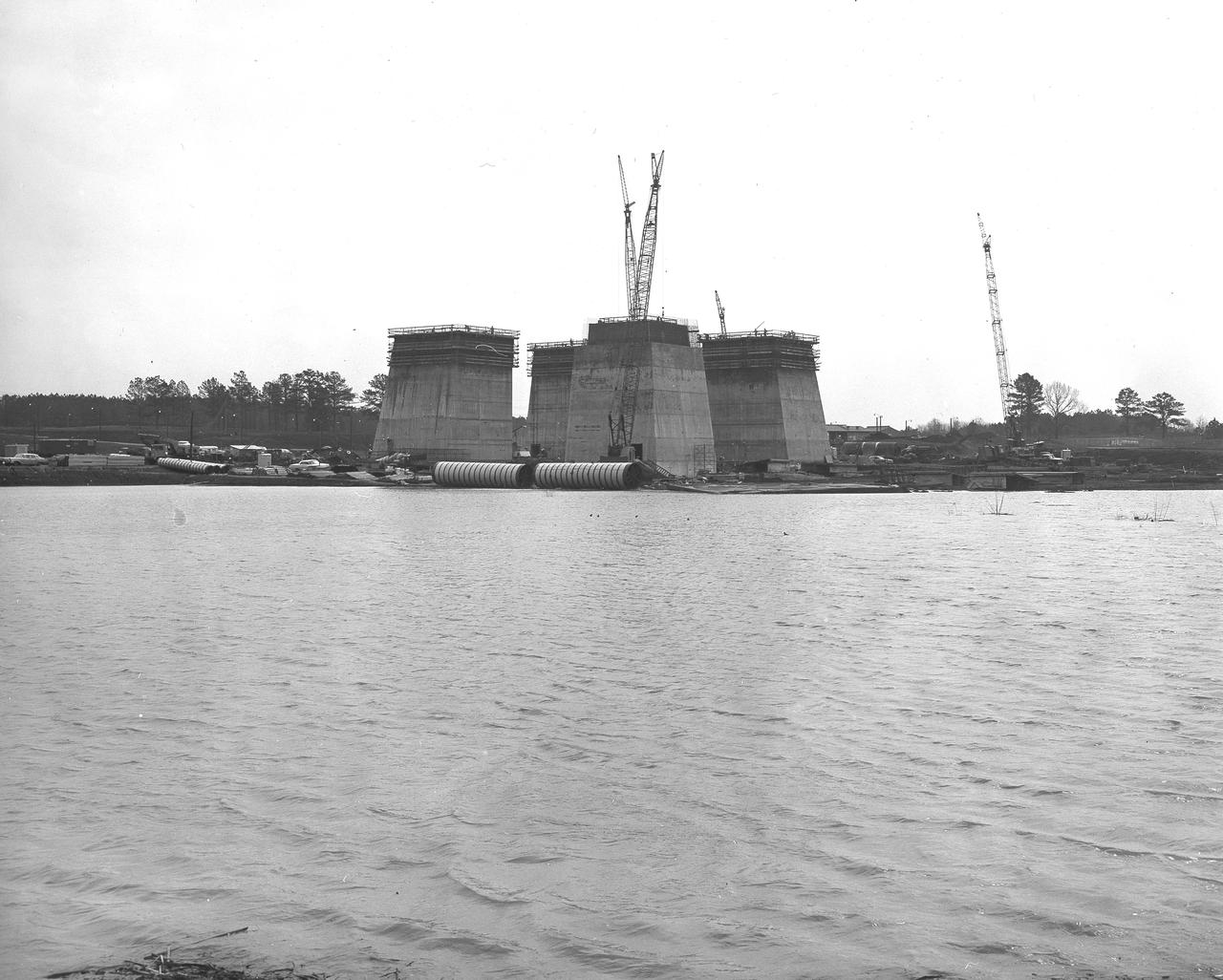 At its founding, the Marshall Space Flight Center (MSFC) inherited the Army’s Jupiter and Redstone test stands, but much larger facilities were needed for the giant stages of the Saturn V. From 1960 to 1964, the existing stands were remodeled and a sizable new test area was developed. The new comprehensive test complex for propulsion and structural dynamics was unique within the nation and the free world, and they remain so today because they were constructed with foresight to meet the future as well as on going needs. Construction of the S-IC Static test stand complex began in 1961 in the west test area of MSFC, and was completed in 1964. The S-IC static test stand was designed to develop and test the 138-ft long and 33-ft diameter Saturn V S-IC first stage, or booster stage, weighing in at 280,000 pounds. Required to hold down the brute force of a 7,500,000-pound thrust produced by 5 F-1 engines, the S-IC static test stand was designed and constructed with the strength of hundreds of tons of steel and 12,000,000 pounds of cement, planted down to bedrock 40 feet below ground level. The foundation walls, constructed with concrete and steel, are 4 feet thick. The base structure consists of four towers with 40-foot-thick walls extending upward 144 feet above ground level. The structure was topped by a crane with a 135-foot boom. With the boom in the upright position, the stand was given an overall height of 405 feet, placing it among the highest structures in Alabama at the time. In the early stages of excavation, a natural spring was disturbed that caused a water problem which required constant pumping from the site and is even pumped to this day. Behind this reservoir of pumped water is the S-IC test stand boasting its ever-growing four towers as of March 29, 1963. 