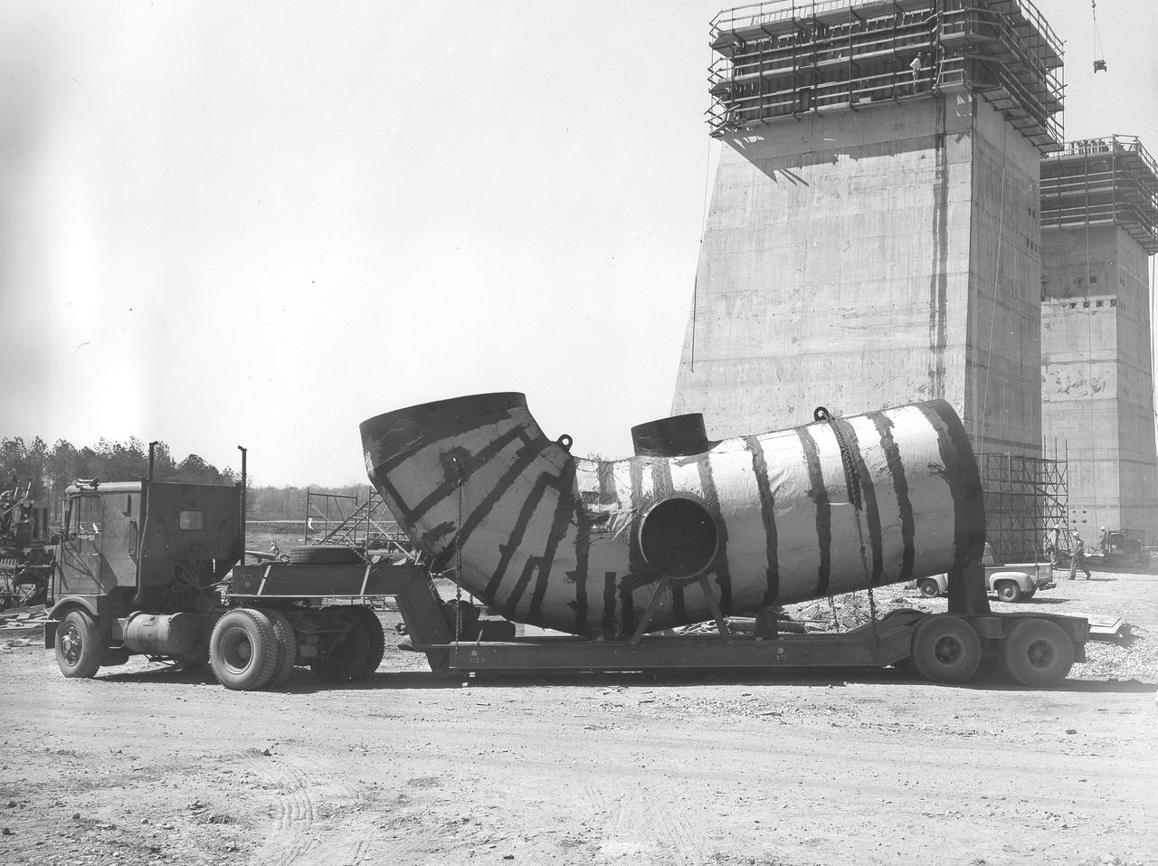 At its founding, the Marshall Space Flight Center (MSFC) inherited the Army’s Jupiter and Redstone test stands, but much larger facilities were needed for the giant stages of the Saturn V. From 1960 to 1964, the existing stands were remodeled and a sizable new test area was developed. The new comprehensive test complex for propulsion and structural dynamics was unique within the nation and the free world, and they remain so today because they were constructed with foresight to meet the future as well as on going needs. Construction of the S-IC Static test stand complex began in 1961 in the west test area of MSFC, and was completed in 1964. The S-IC static test stand was designed to develop and test the 138-ft long and 33-ft diameter Saturn V S-IC first stage, or booster stage, weighing in at 280,000 pounds. Required to hold down the brute force of a 7,500,000-pound thrust produced by 5 F-1 engines, the S-IC static test stand was designed and constructed with the strength of hundreds of tons of steel and 12,000,000 pounds of cement, planted down to bedrock 40 feet below ground level. The foundation walls, constructed with concrete and steel, are 4 feet thick. The base structure consists of four towers with 40-foot-thick walls extending upward 144 feet above ground level. The structure was topped by a crane with a 135-foot boom. With the boom in the upright position, the stand was given an overall height of 405 feet, placing it among the highest structures in Alabama at the time. In addition to the stand itself, related facilities were constructed during this time. Built directly east of the test stand was the Block House, which served as the control center for the test stand. The two were connected by a narrow access tunnel which housed the cables for the controls. Again to the east, just south of the Block House, was a newly constructed Pump House. Its function was to provide water to the stand to prevent melting damage during testing. The water was sprayed through small holes in the stand’s 1900 ton water deflector at the rate of 320,000 gallons per minute. This photo, taken April 4, 1963, depicts the portion of the massive water line that was installed into the S-IC test Stand.