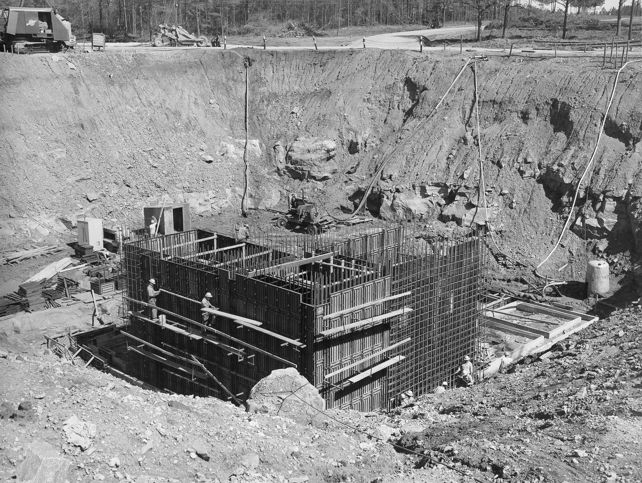 At its founding, the Marshall Space Flight Center (MSFC) inherited the Army’s Jupiter and Redstone test stands, but much larger facilities were needed for the giant stages of the Saturn V. From 1960 to 1964, the existing stands were remodeled and a sizable new test area was developed. The new comprehensive test complex for propulsion and structural dynamics was unique within the nation and the free world, and they remain so today because they were constructed with foresight to meet the future as well as on going needs. Construction of the S-IC Static test stand complex began in 1961 in the west test area of MSFC, and was completed in 1964. The S-IC static test stand was designed to develop and test the 138-ft long and 33-ft diameter Saturn V S-IC first stage, or booster stage, weighing in at 280,000 pounds. Required to hold down the brute force of a 7,500,000-pound thrust produced by 5 F-1 engines, the S-IC static test stand was designed and constructed with the strength of hundreds of tons of steel and 12,000,000 pounds of cement, planted down to bedrock 40 feet below ground level. The foundation walls, constructed with concrete and steel, are 4 feet thick. The base structure consists of four towers with 40-foot-thick walls extending upward 144 feet above ground level. The structure was topped by a crane with a 135-foot boom. With the boom in the upright position, the stand was given an overall height of 405 feet, placing it among the highest structures in Alabama at the time. North of the massive S-IC test stand, the F-1 Engine test stand was built. Designed to assist in the development of the F-1 Engine, the F-1 test stand is a vertical engine firing test stand, 239 feet in elevation and 4,600 square feet in area at the base. Capability was provided for static firing of 1.5 million pounds of thrust using liquid oxygen and kerosene. Like the S-IC stand, the foundation of the F-1 stand is keyed into the bedrock approximately 40 feet below grade. This photo, taken April 4, 1963 depicts the construction of the F-1 test stand foundation walls.
